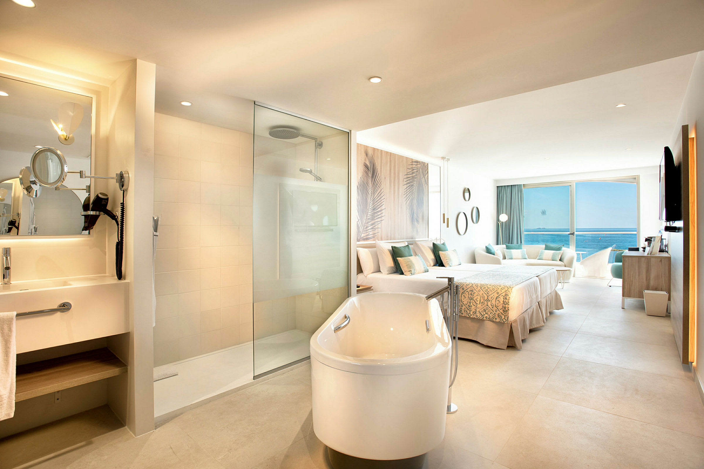 Hotellrum med öppen planlösning som inkluderar ett badkar, dusch, dubbelsäng och sittgrupp med utsikt över havet. (AI-genererad text)