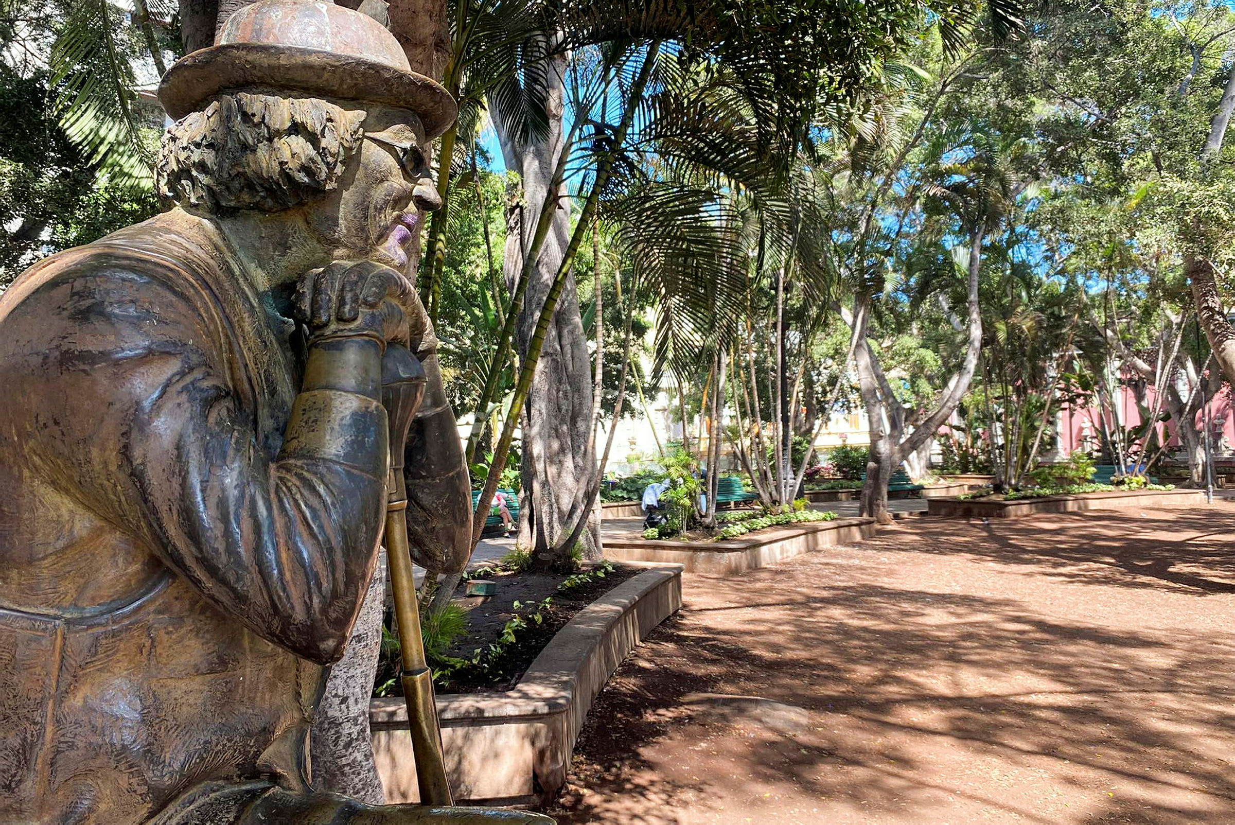 En staty av en person med hatt och glasögon, placerad i en grönskande park. (AI-genererad text)