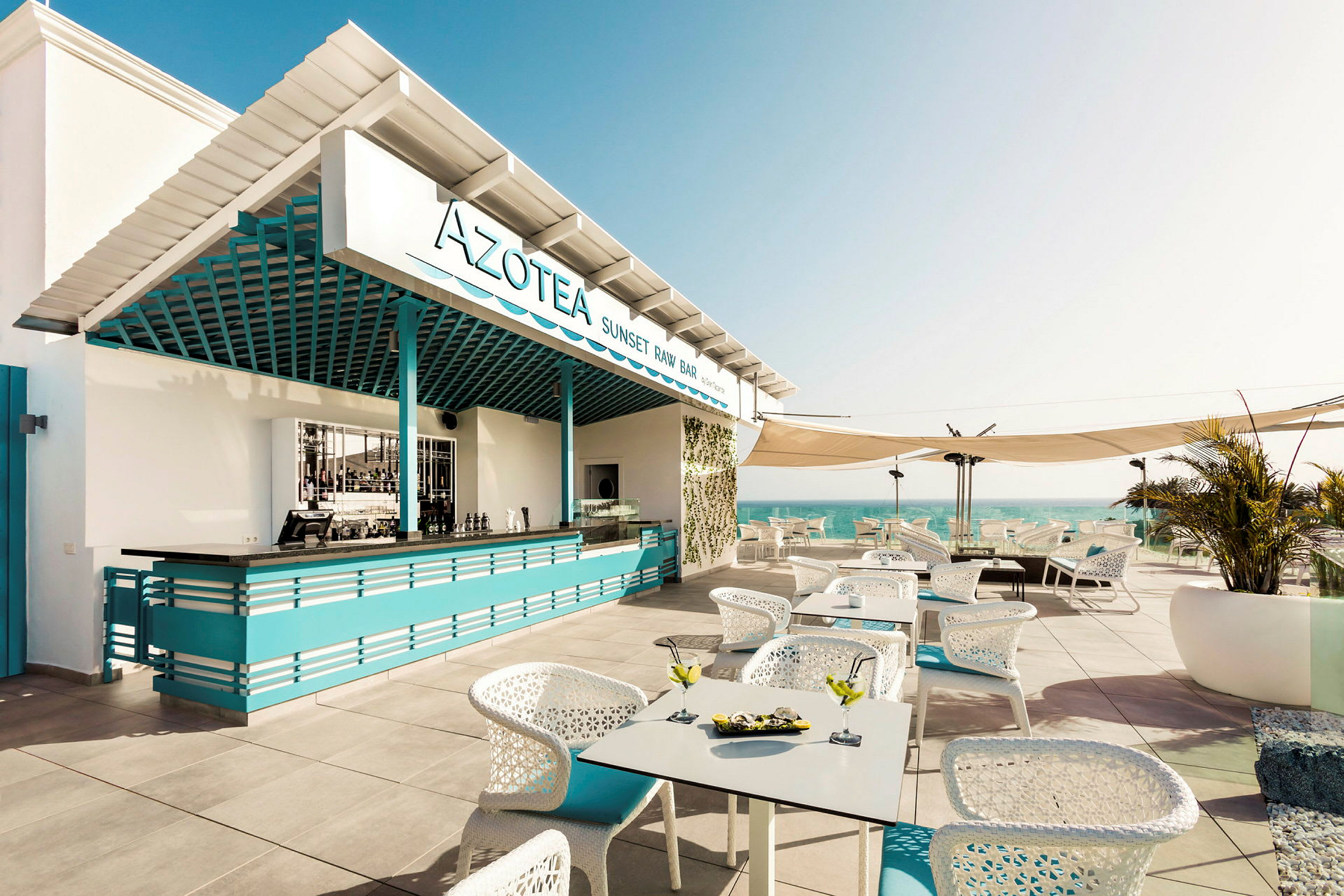 En öppen terrass med en bar skyltad som Azotea Sunset Raw Bar, sittplatser och utsikt över havet. (AI-genererad text)