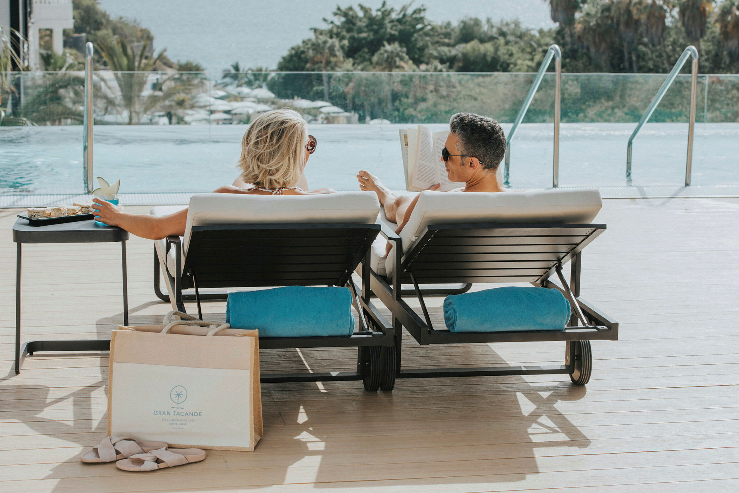 Två personer sitter på solstolar vid en pool med utsikt över havet. (AI-genererad text)