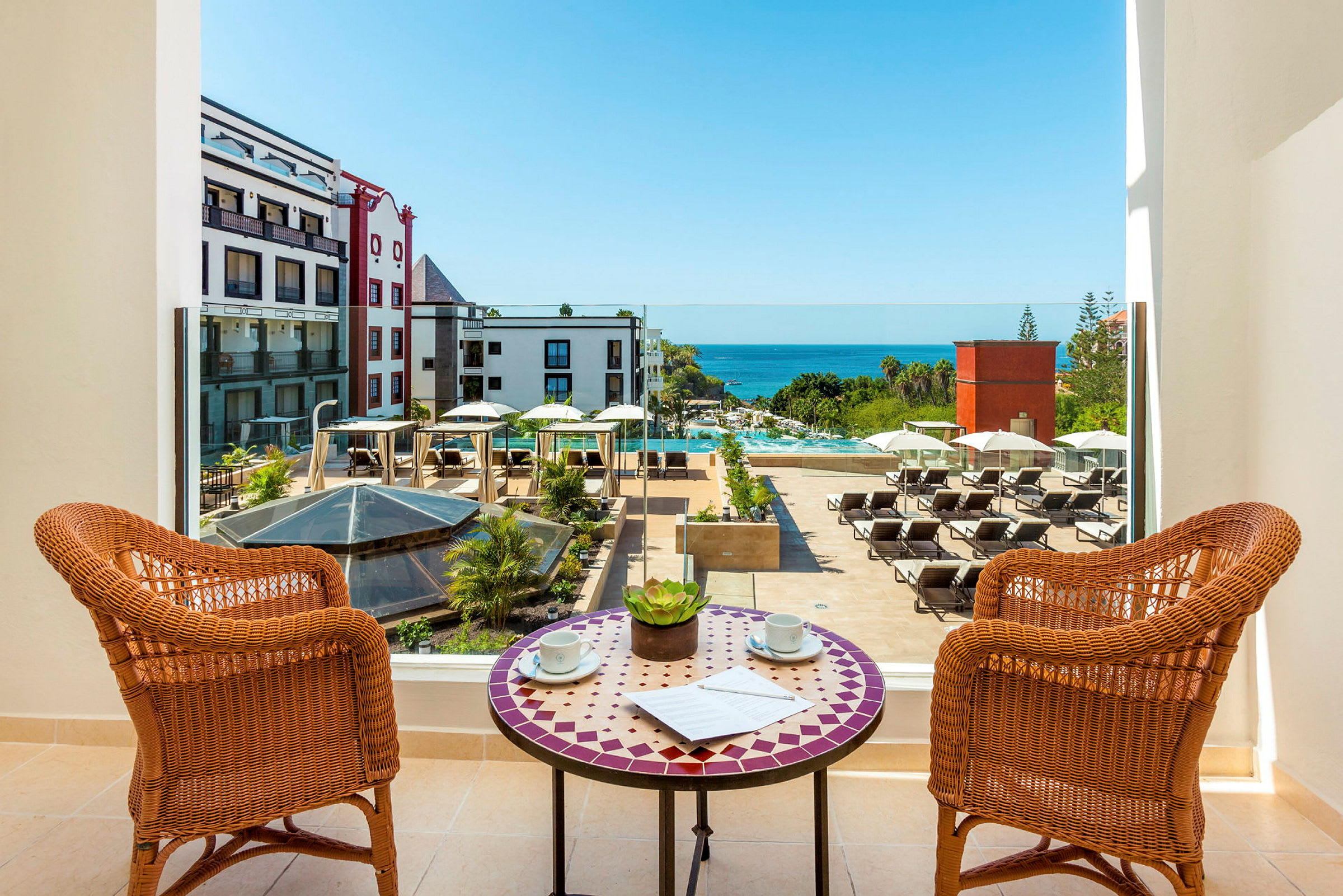 Utsikt från en balkong över en hotellpool med solstolar och havet i bakgrunden. (AI-genererad text)
