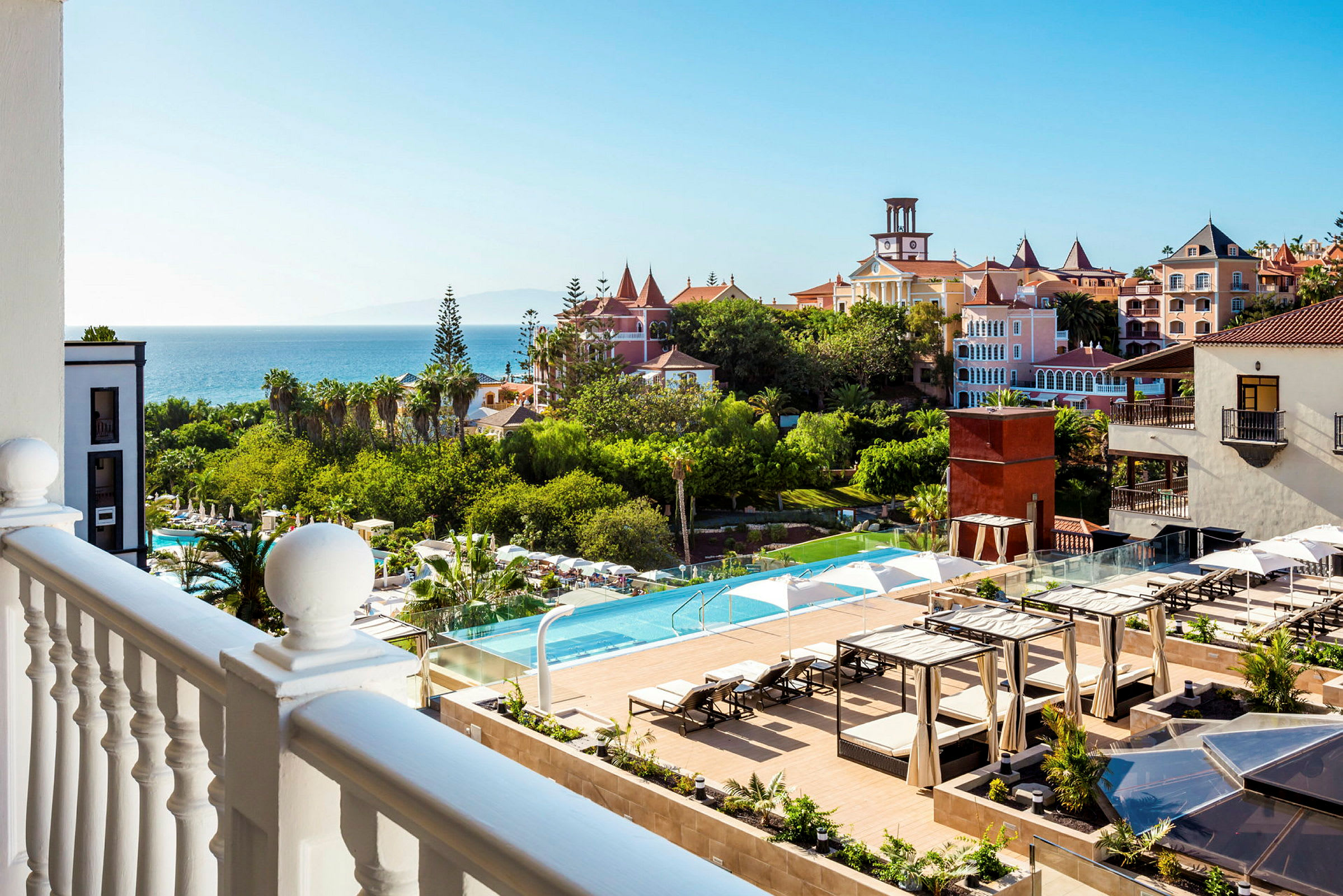 Utsikt från balkong över hotellområdet med pool och trädgård samt havet i bakgrunden. (AI-genererad text)