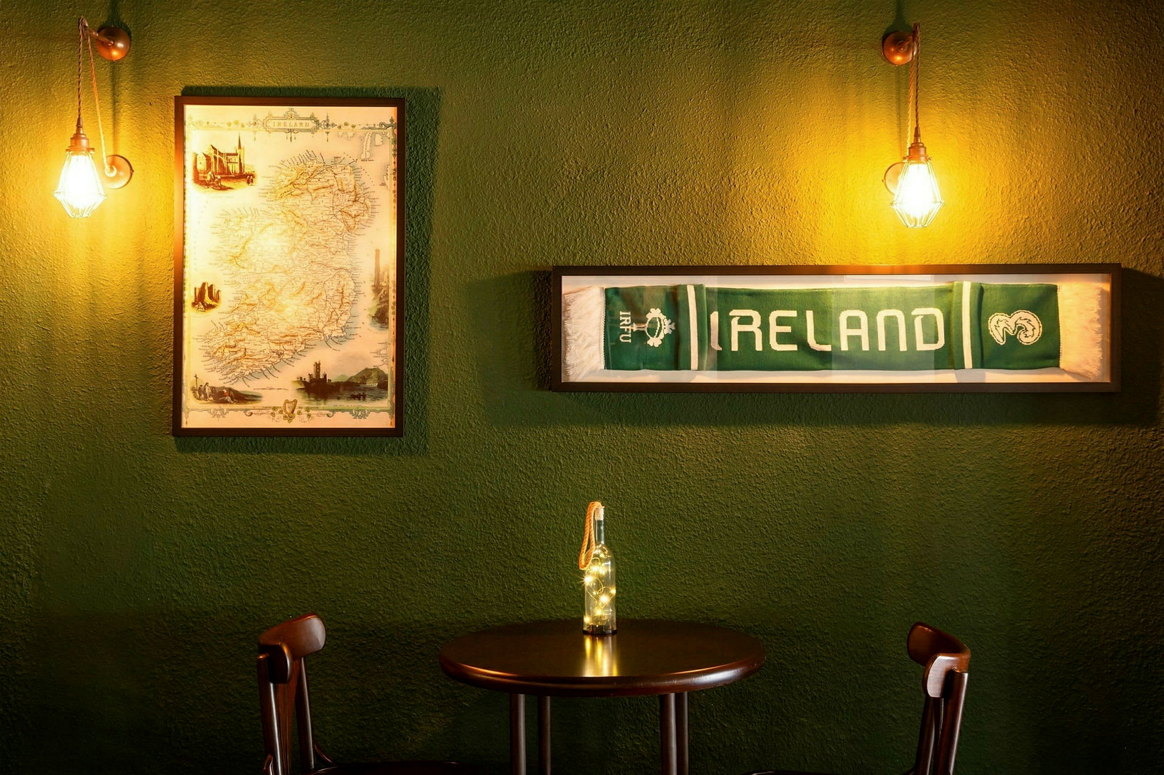 Ett bord med två stolar framför en grön vägg, dekorerad med en inramad karta över Irland och en inramad halsduk med texten "IRELAND". (AI-genererad text)