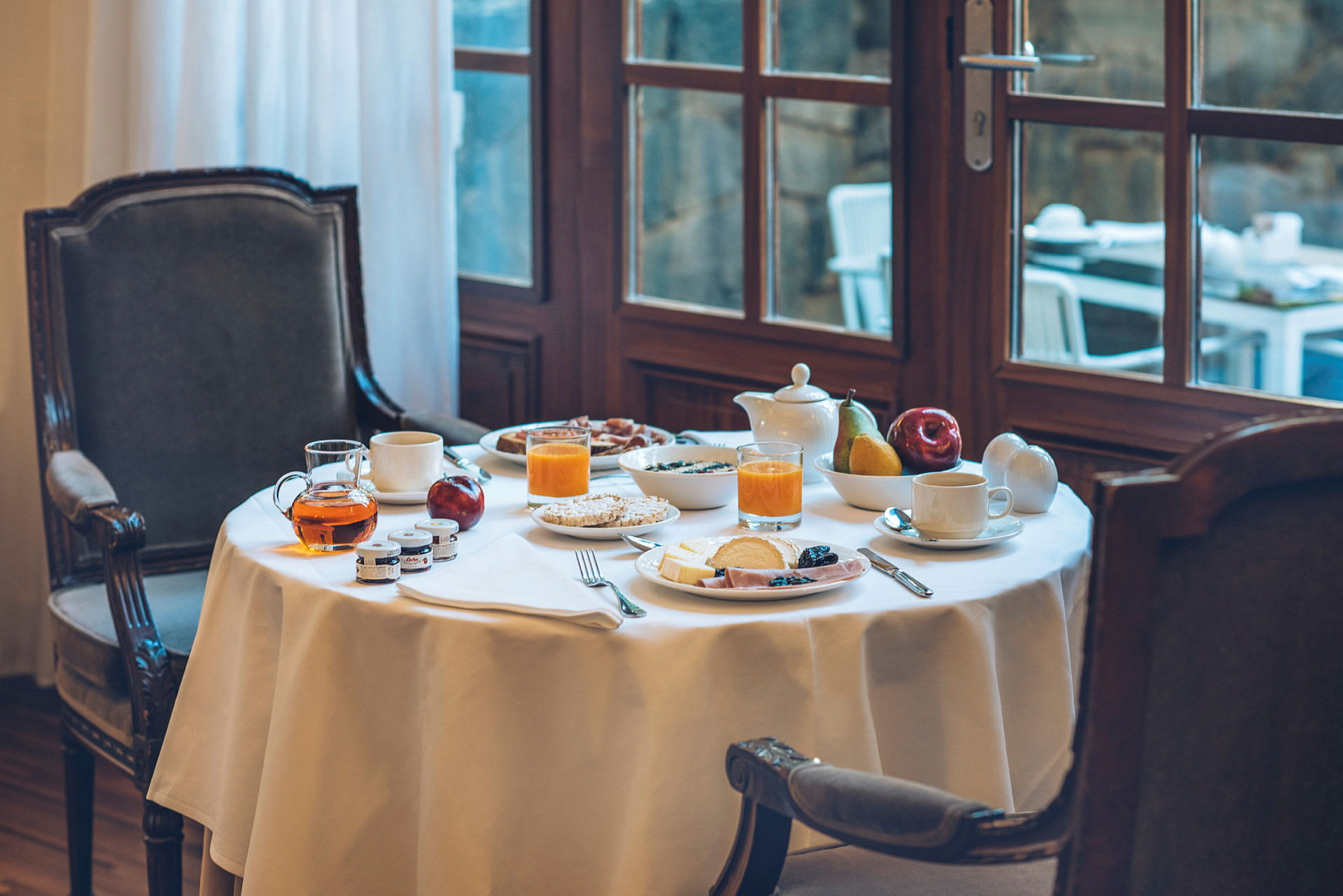 Ett dukat frukostbord med glas apelsinjuice, te, frukt och skivade pålägg. Bordet står vid ett fönster. (AI-genererad text)