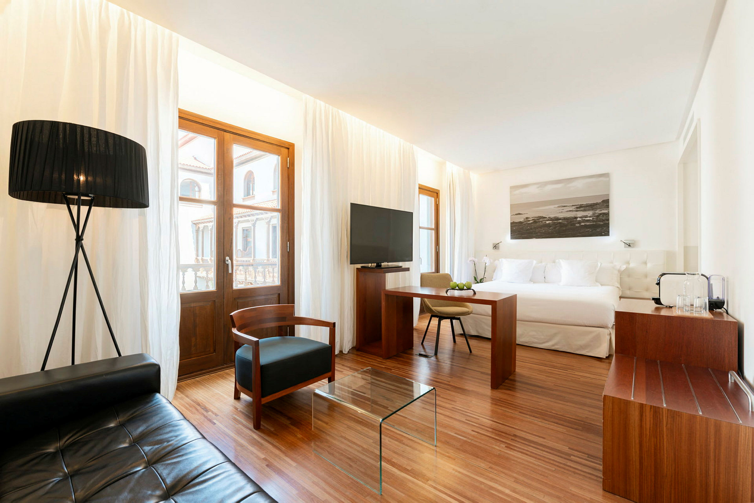 Ett modernt inrett hotellrum med en stor säng, sittgrupp med textilmöbler och en glasbord, samt fönster med vita gardiner. (AI-genererad text)