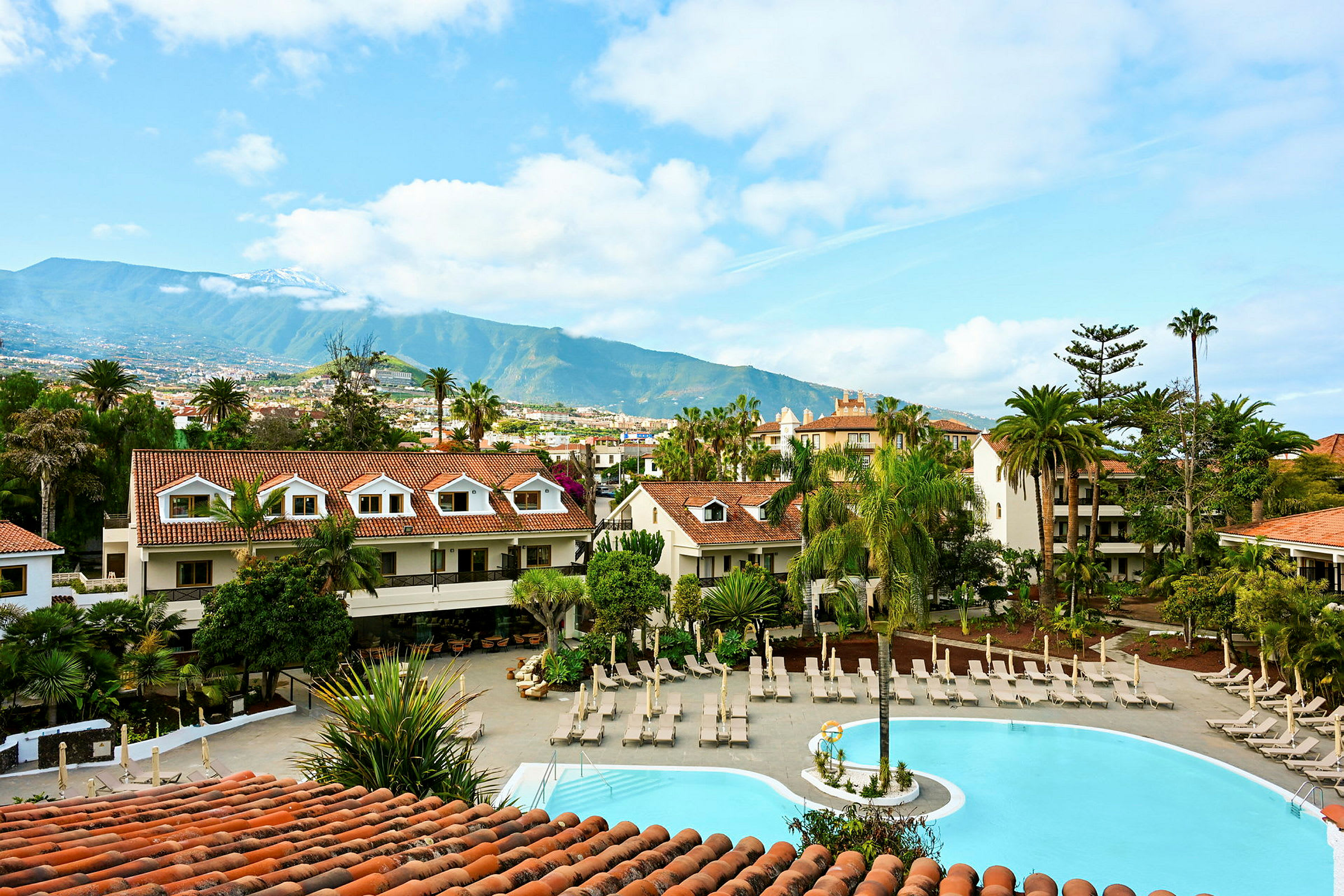 Ett hotellområde med en pool omgiven av solstolar och grönska med berg i bakgrunden. (AI-genererad text)