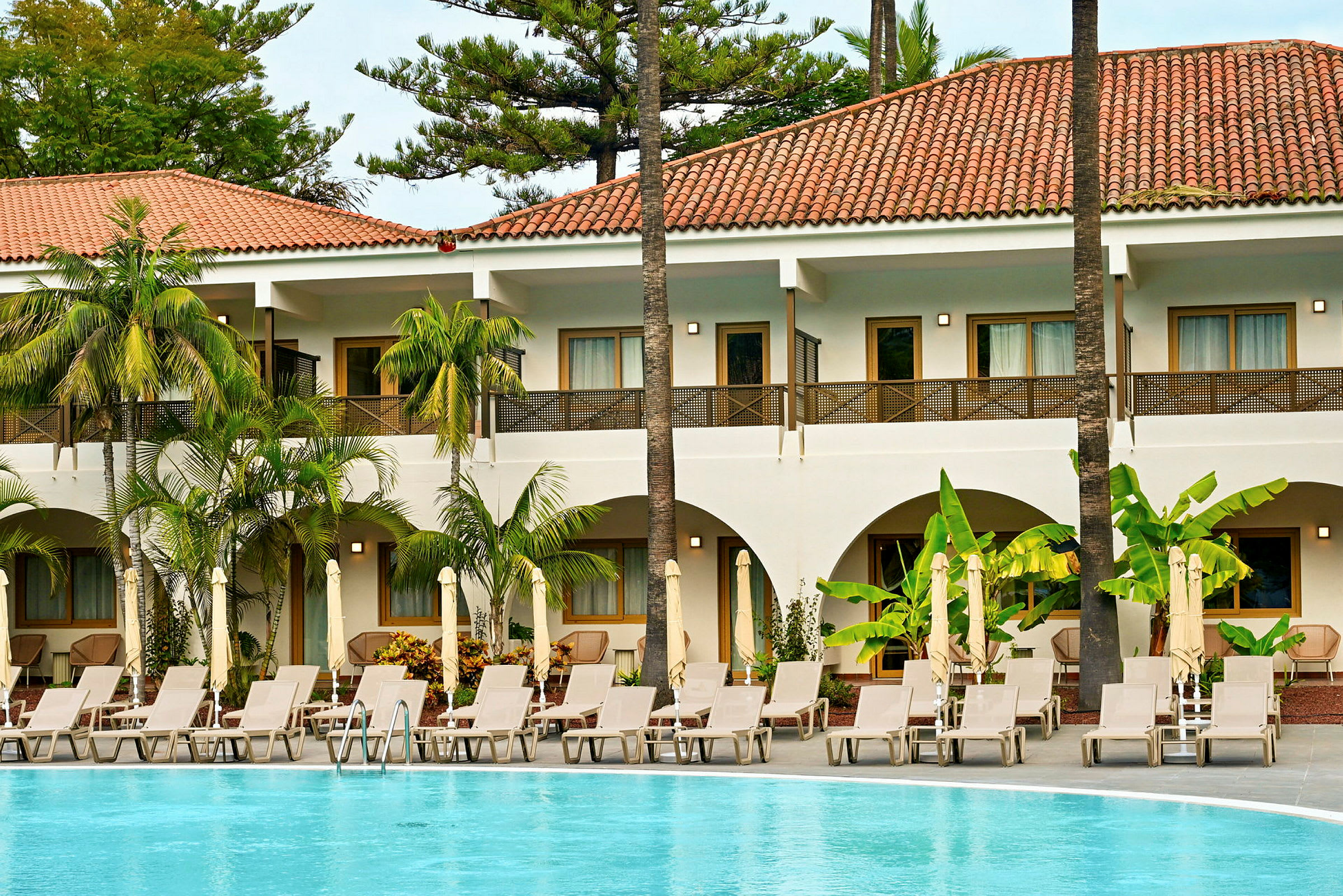 Tvåvåningshotell med pool, solstolar och gröna växter i omgivningen. (AI-genererad text)
