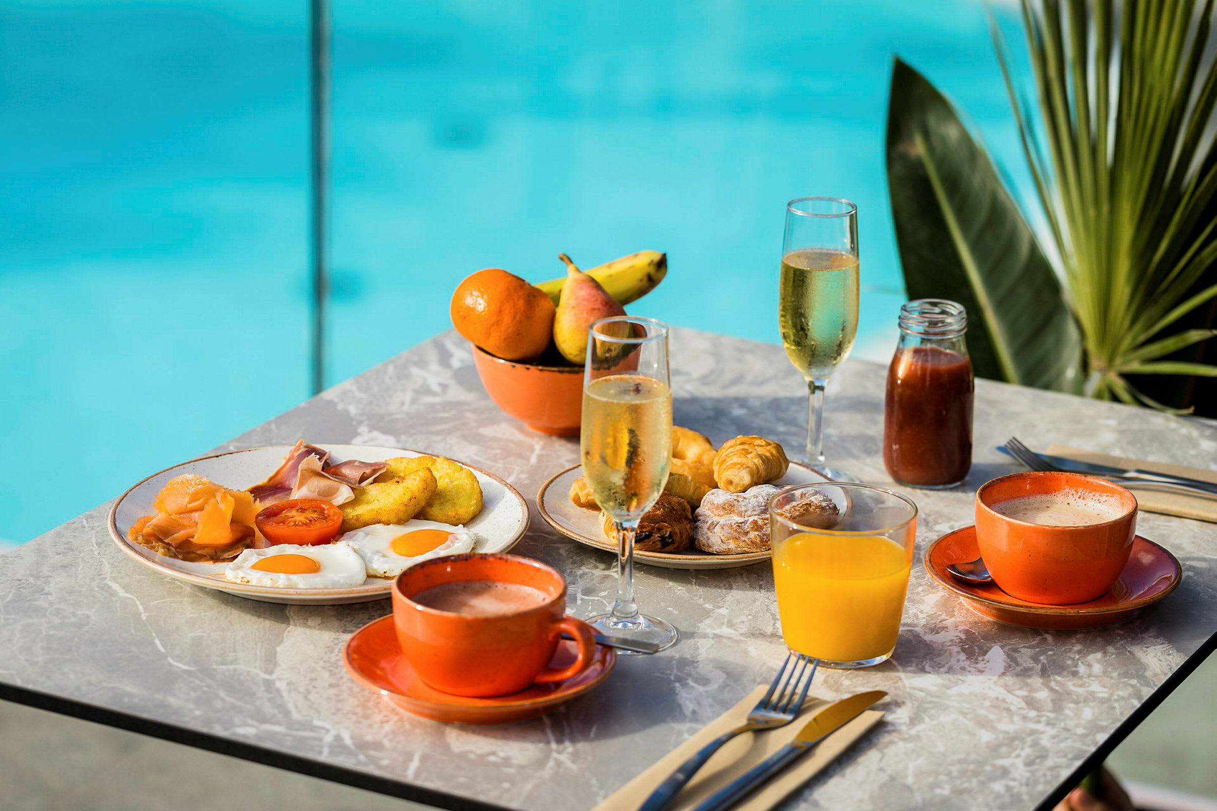 Frukostbord med diverse mat såsom ägg, korv, croissanter och drycker som juice och kaffe. En pool syns i bakgrunden. (AI-genererad text)