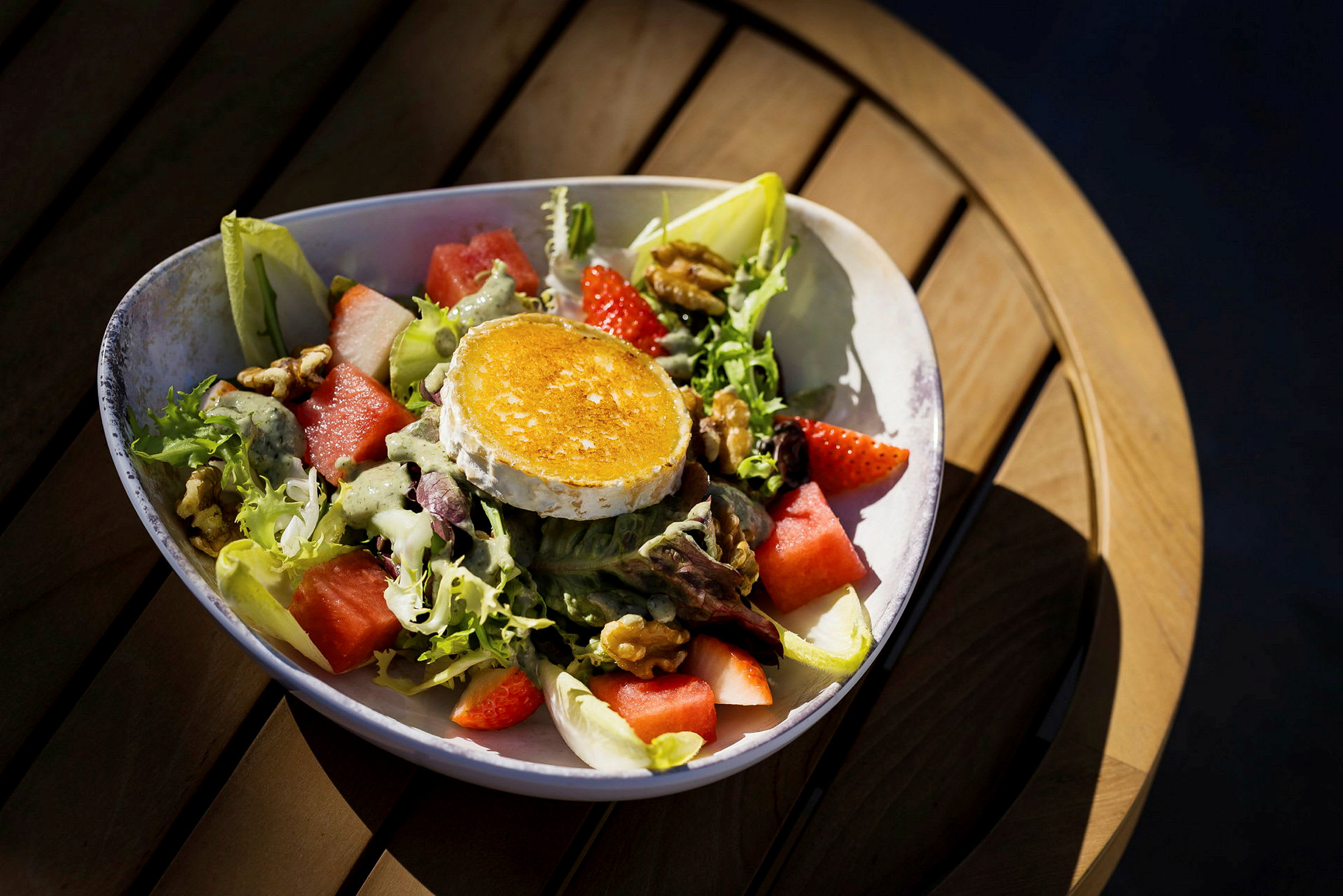 En tallrik med en sallad som innehåller grillad ostskiva, melon, jordgubbar, nötter och salladsblad. Tallriken står på ett träbord. (AI-genererad text)