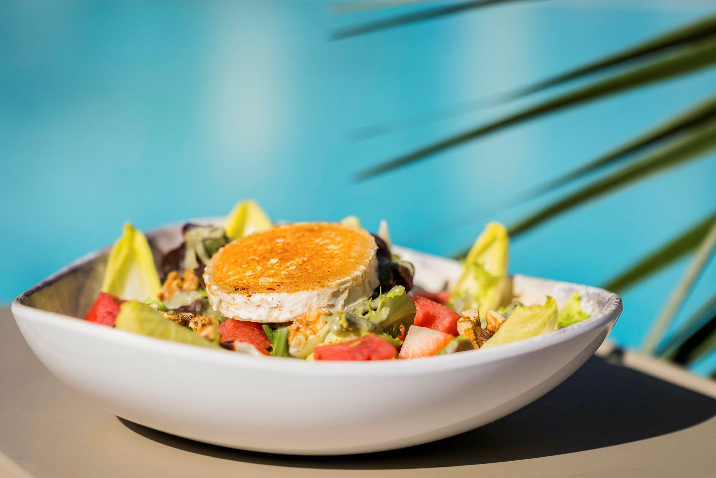 En sallad med olika grönsaker och en bit gratinerad ost serverad i en vit skål, med suddig bakgrund av blått vatten och gröna blad. (AI-genererad text)