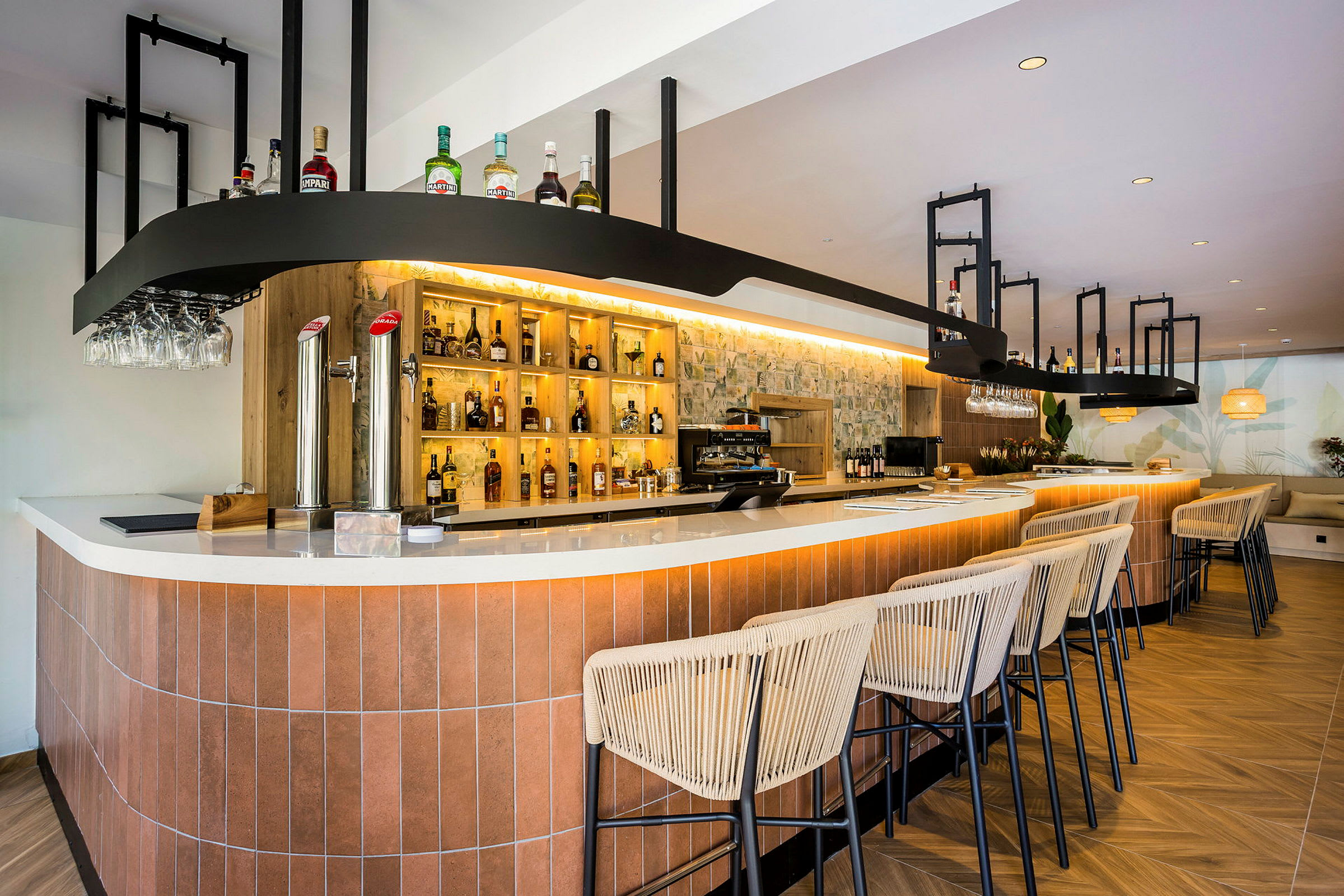 En modern bar med en lång bardisk, upplysta hyllor med flaskor och bekväma barstolar i trä och linne. (AI-genererad text)