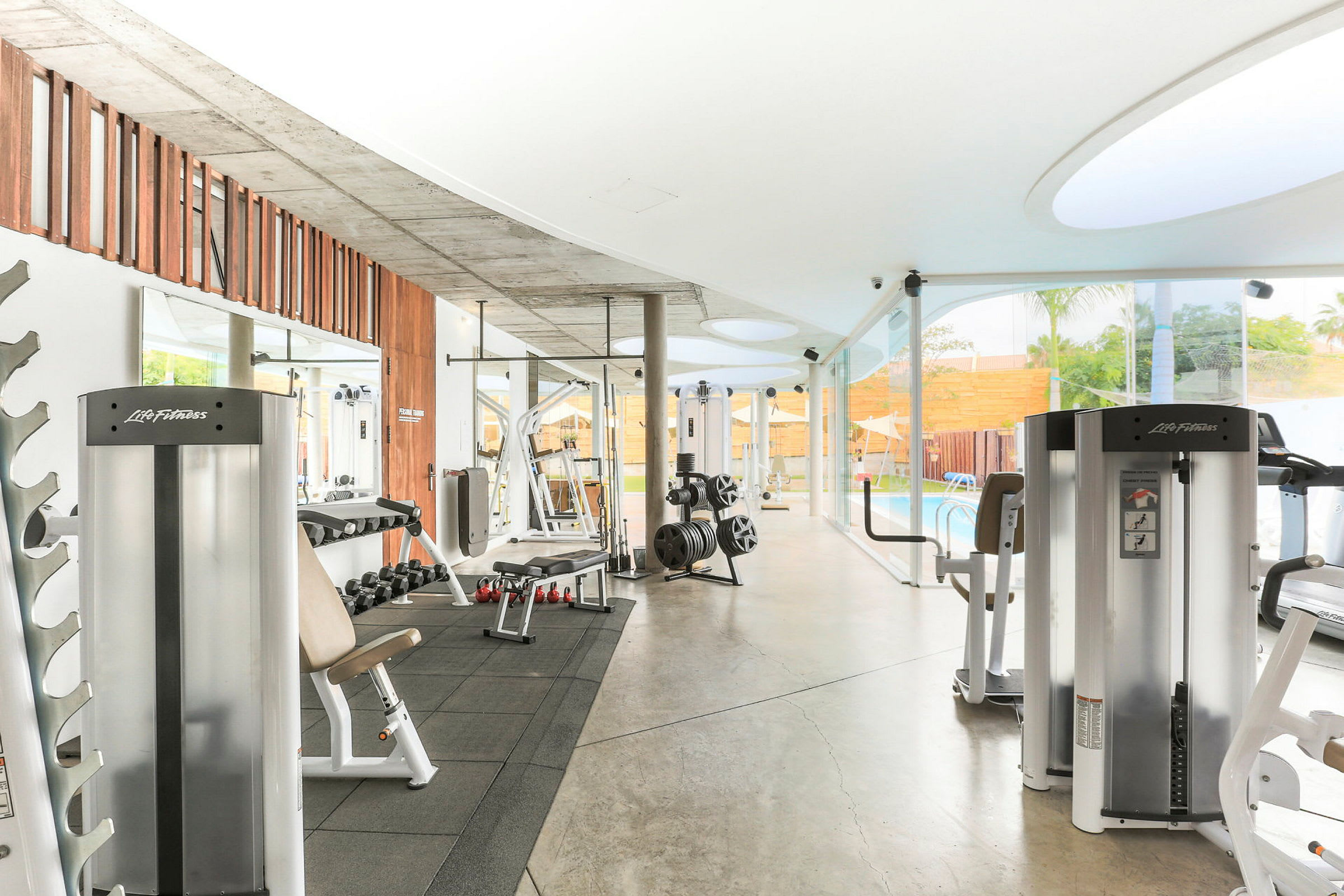 Ett modernt gym med träningsmaskiner och fria vikter, beläget nära ett glasparti med utsikt över en pool. (AI-genererad text)
