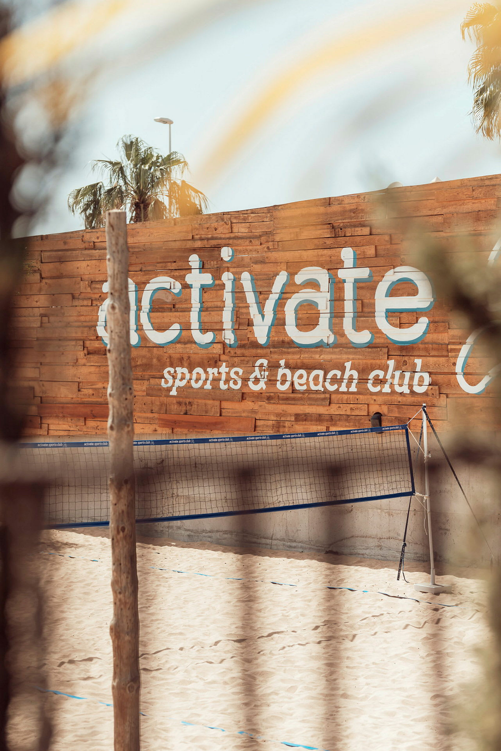 Sandstrand med volleybollnät framför en trävägg med texten 'activate sports & beach club'. (AI-genererad text)