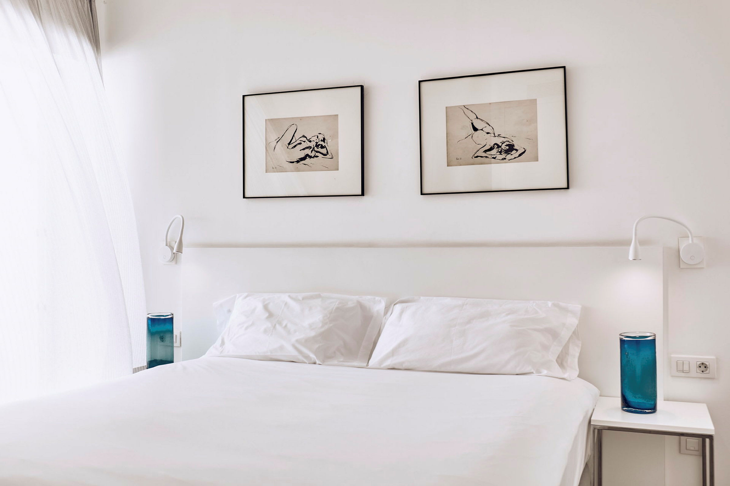 Ett hotellrum med en vit dubbelsäng, två inramade konstverk på väggen och blå dekorativa föremål på nattygsborden. (AI-genererad text)