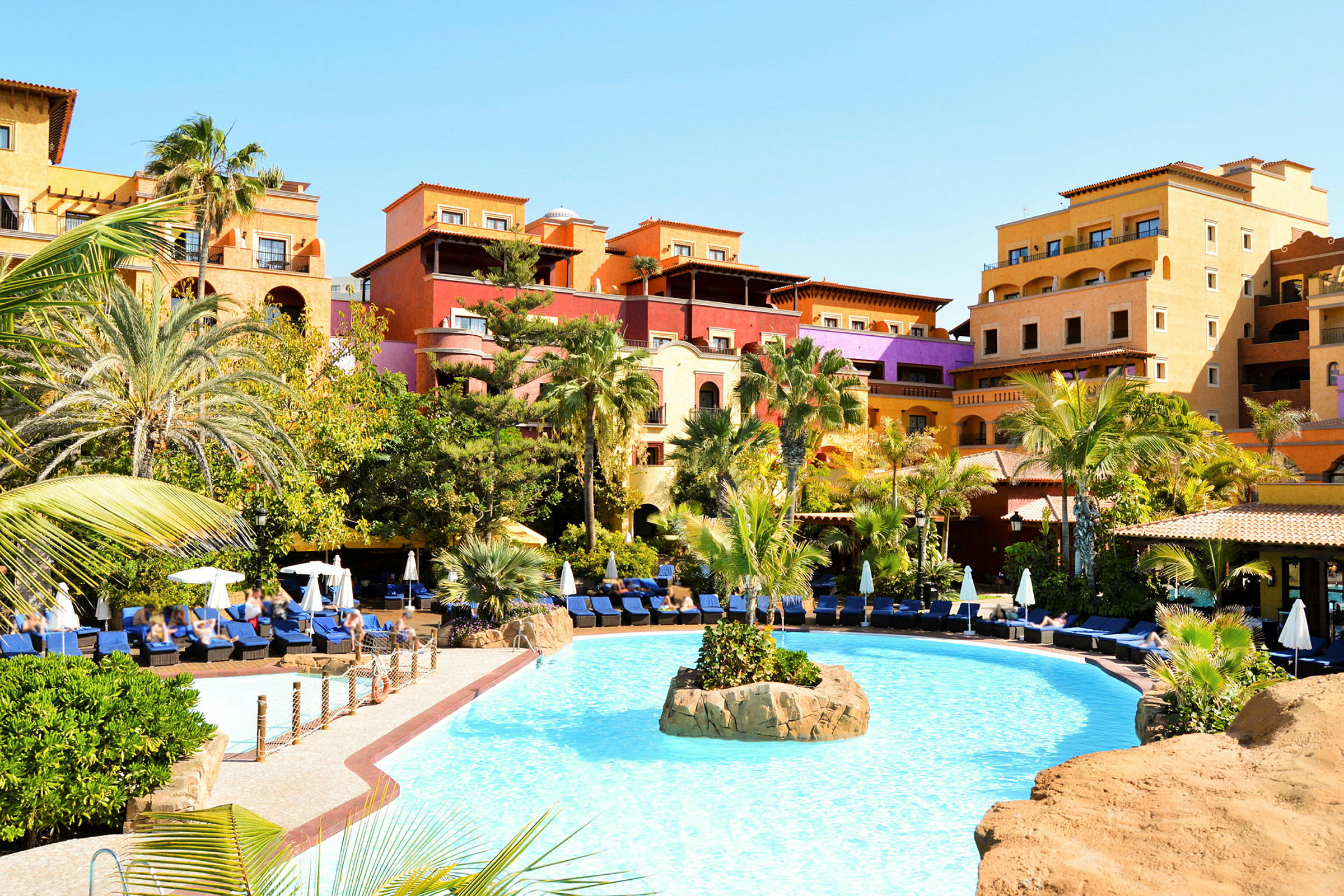 Hotellbyggnad med starka färger i bakgrunden, framför en pool och solstolar omgivna av palmer och grönska. (AI-genererad text)