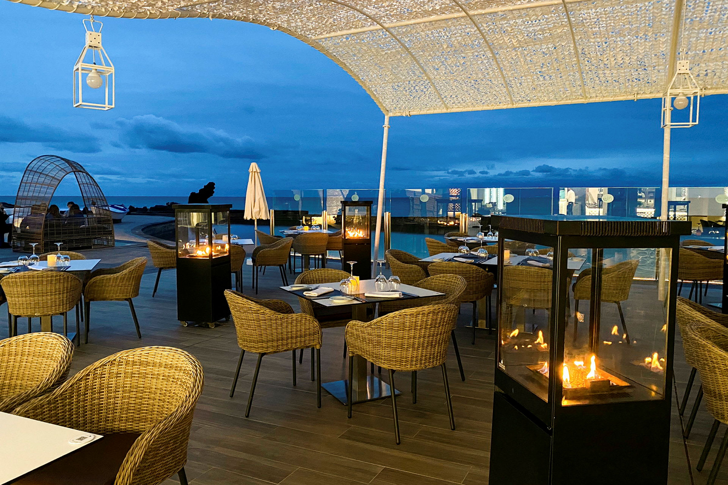 Utomhusrestaurang med bord, stolar och eldstäder under kvällshimlen. (AI-genererad text)