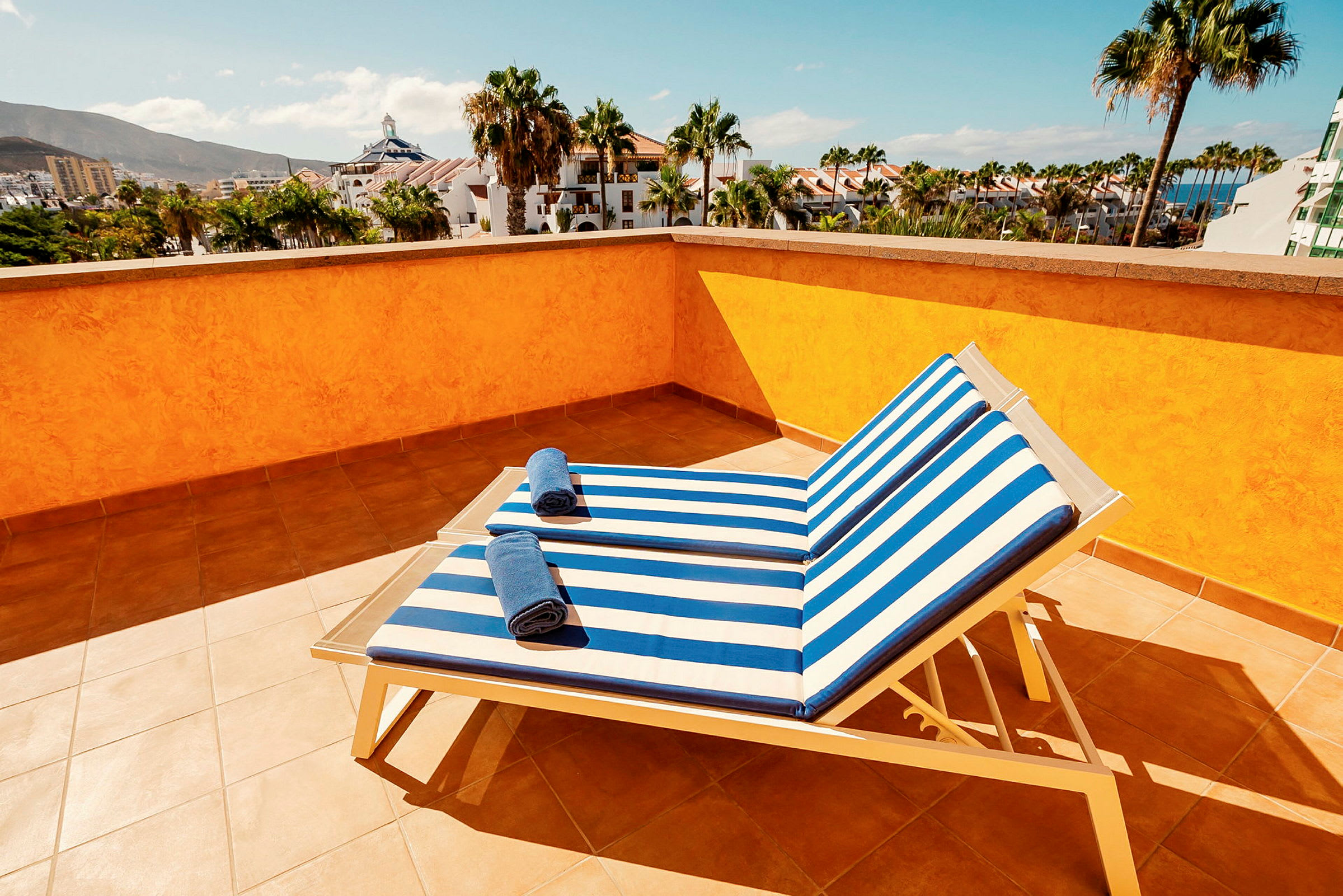 Utsikt från en solterrass med två solstolar med randiga blå och vita dynor samt handdukar på dem, omgivna av palmer och byggnader. (AI-genererad text)