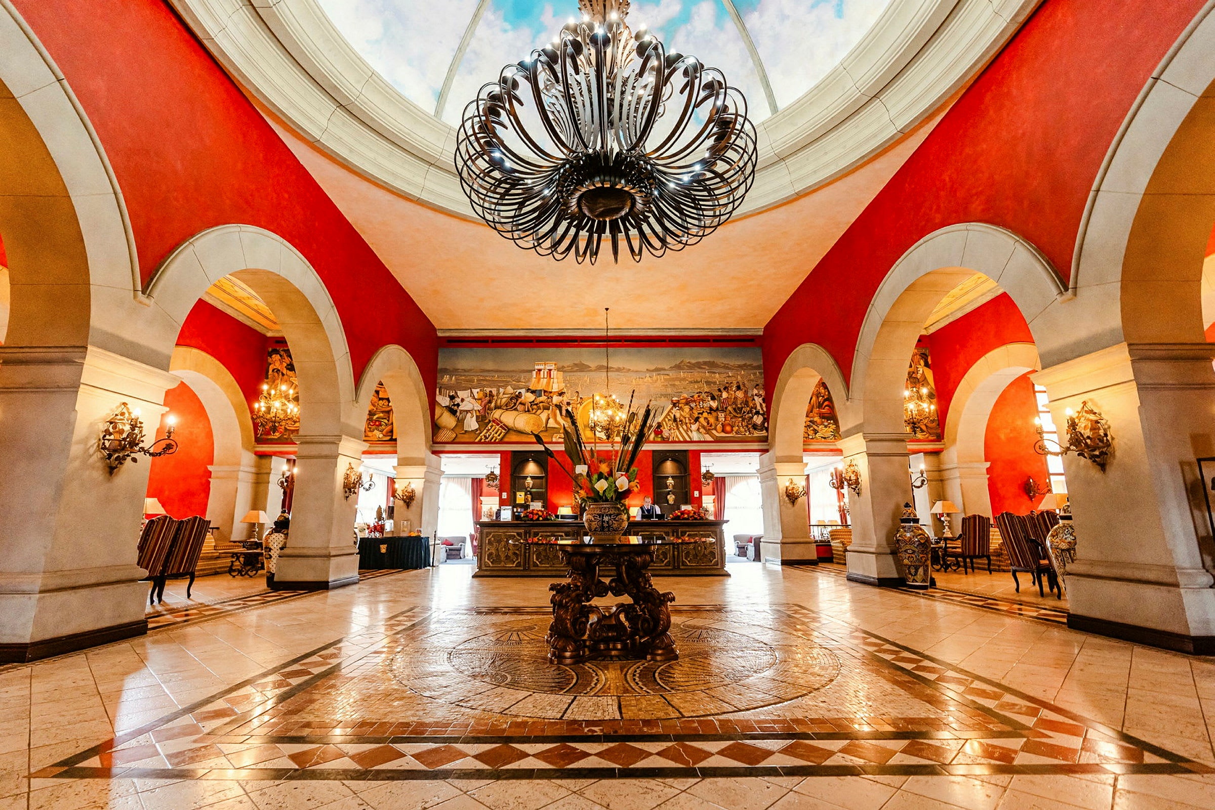 Ett hotellobby med en stor dekorerad ljuskrona i taket, röda väggar med valvbågar och ett mönstrat golv. (AI-genererad text)