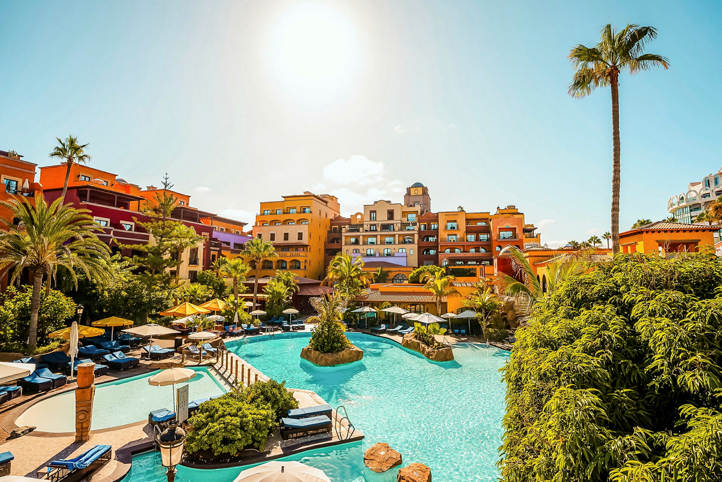 Semesteranläggning med pool, solstolar och färgglada hotellanläggningar under klarblå himmel. (AI-genererad text)