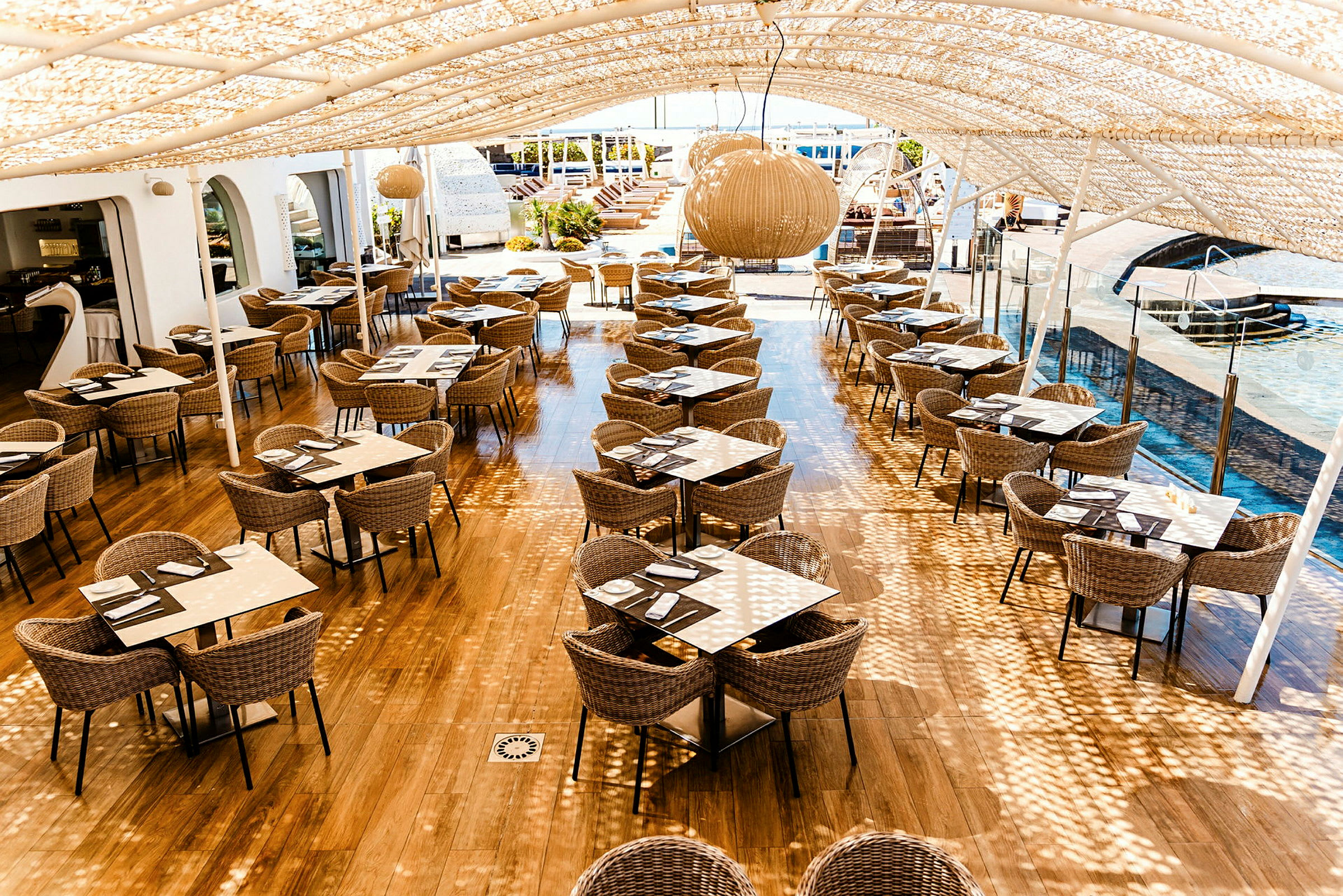 Utomhusrestaurang med takskuggning och sittplatser vid bord av rotting och glas. (AI-genererad text)