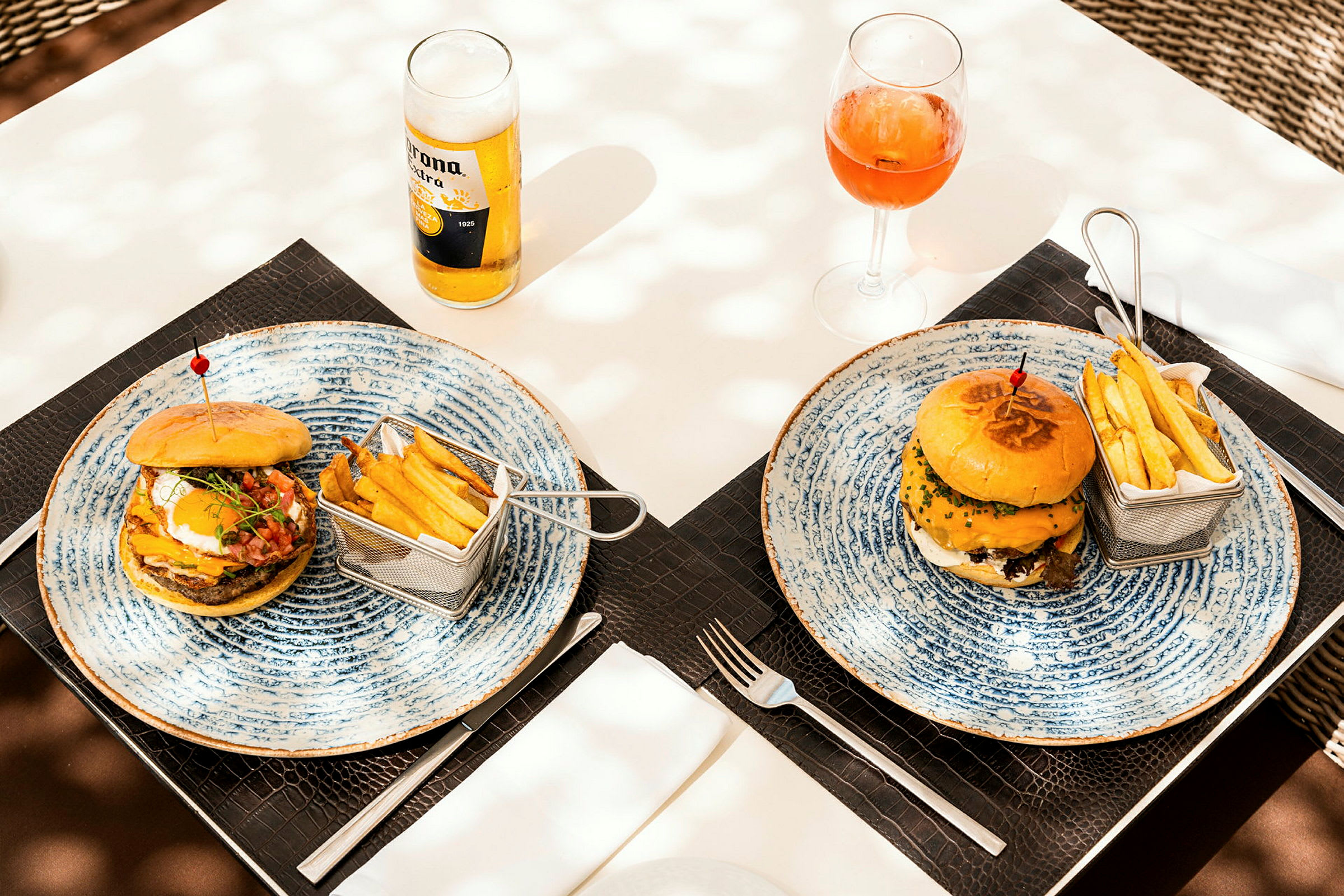 Två tallrikar med hamburgare, pommes frites i små korgar, en öl och ett glas med dryck på ett dukat bord. (AI-genererad text)
