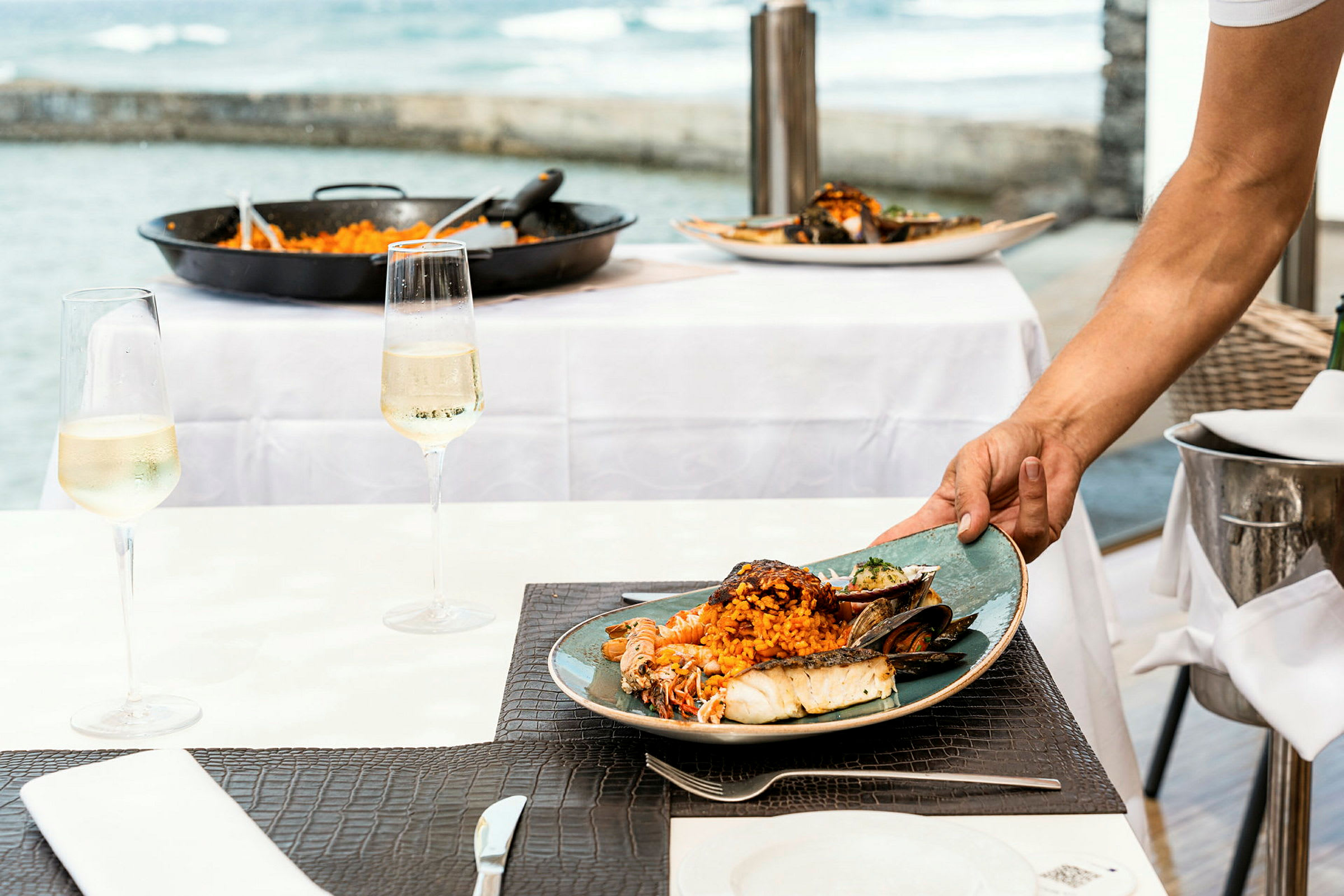 En tallrik med mat, bestående av skaldjur och ris, serveras på ett bord. Bakom bordet är en havsutsikt synlig. Ett glas vin står på bordet. (AI-genererad text)