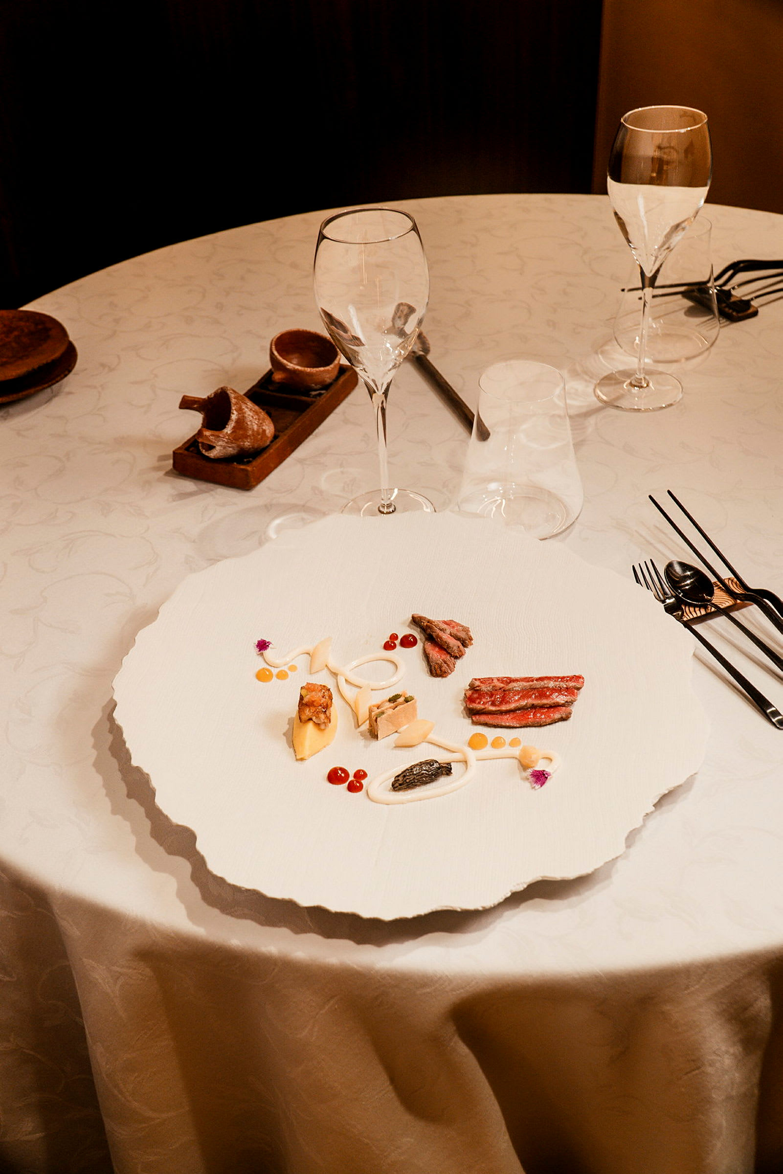 Ett dukat bord med en väldekorerad tallrik innehållande olika små matportioner, omgiven av glas och bestick. (AI-genererad text)