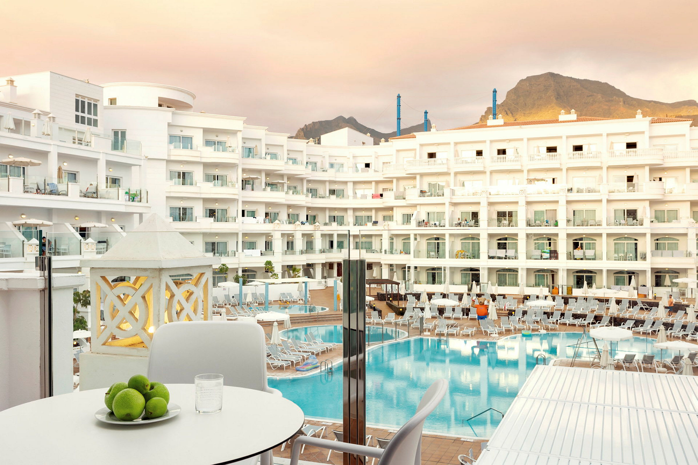 Ett hotellkomplex med flera våningar, en stor pool och solstolar i förgrunden, samt ett bord med fruktskål och glas. (AI-genererad text)