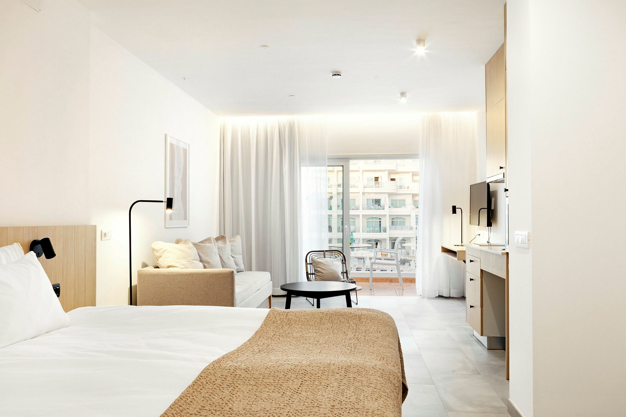 Modern hotellrum med säng, sittgrupp och arbetsyta, belyst av naturligt ljus från balkongdörrar. (AI-genererad text)