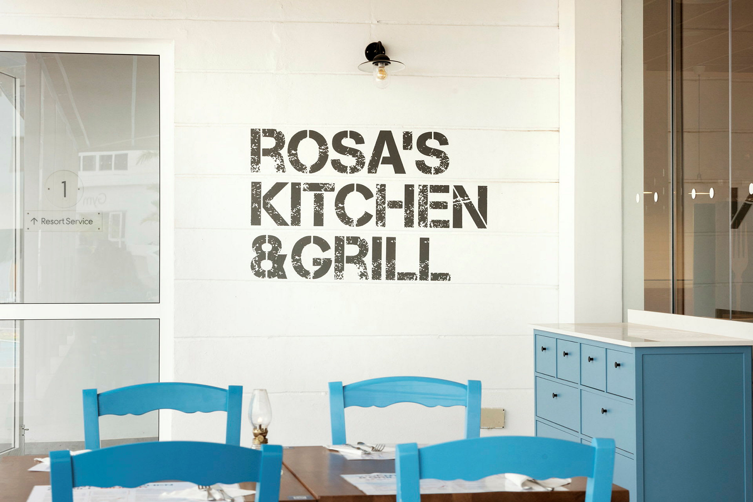 Skylt med texten 'Rosa's Kitchen & Grill' på en vägg, bord och blå stolar i förgrunden. (AI-genererad text)