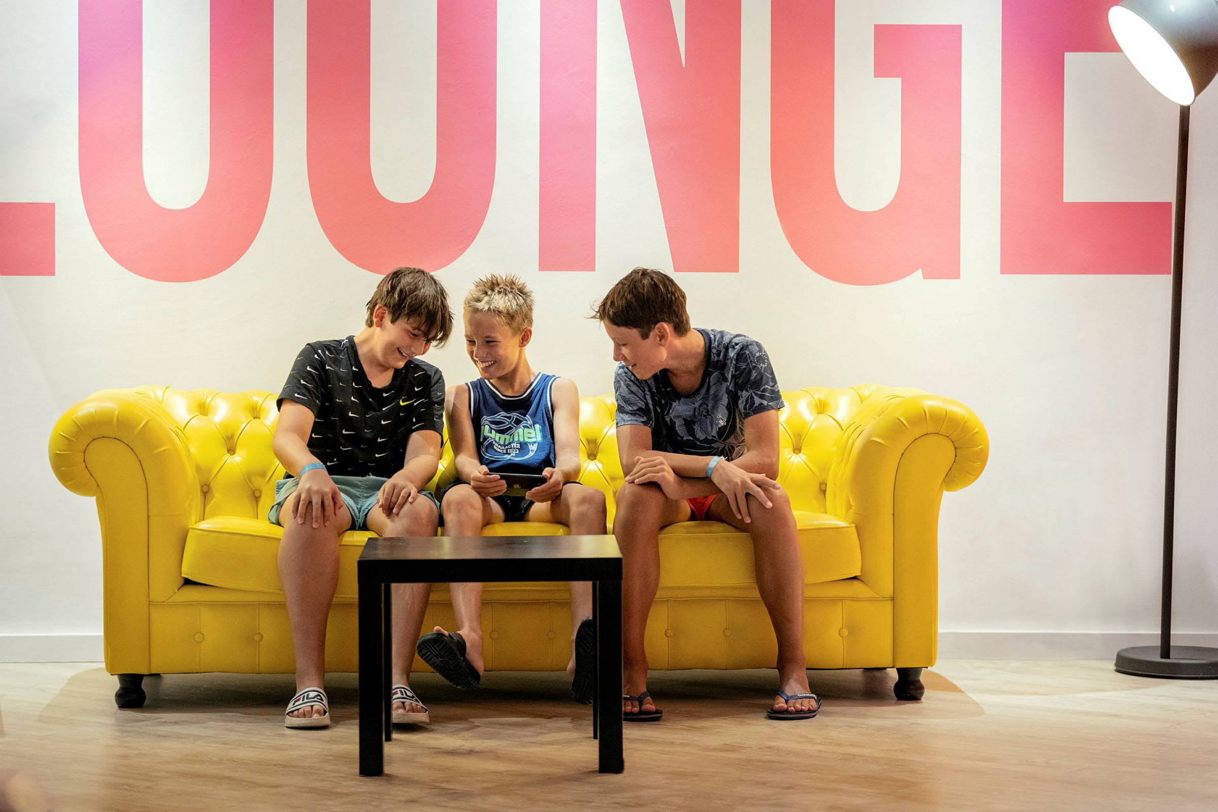 Personer sitter på en gul soffa med ett bord framför, och ordet 'LOUNGE' är synligt på väggen. (AI-genererad text)