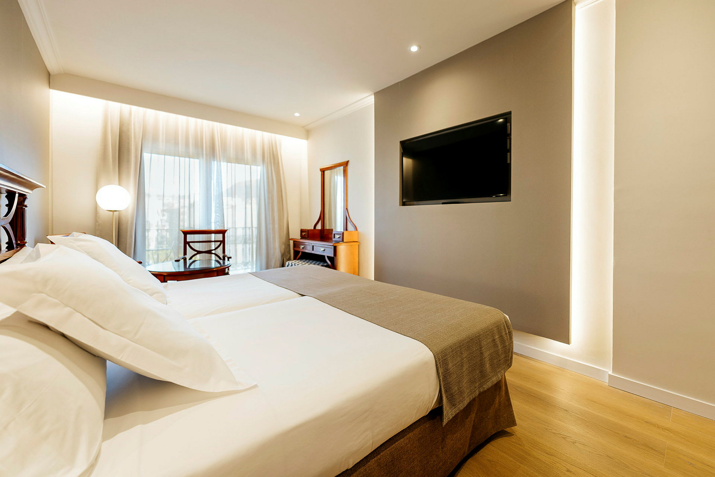 Ett hotellrum med en stor säng, TV på väggen och en byrå framför ett fönster med gardiner. (AI-genererad text)