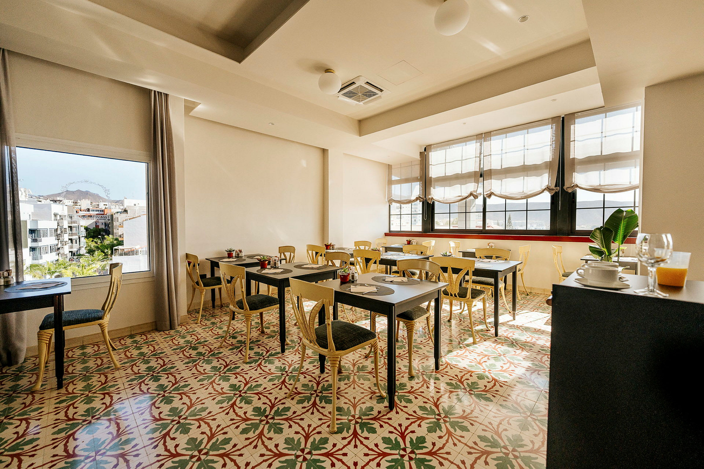 Ett ljust och luftigt café eller restaurang med bord och stolar, en färgglad golvdesign, stora fönster och utsikt över ett stadslandskap. (AI-genererad text)