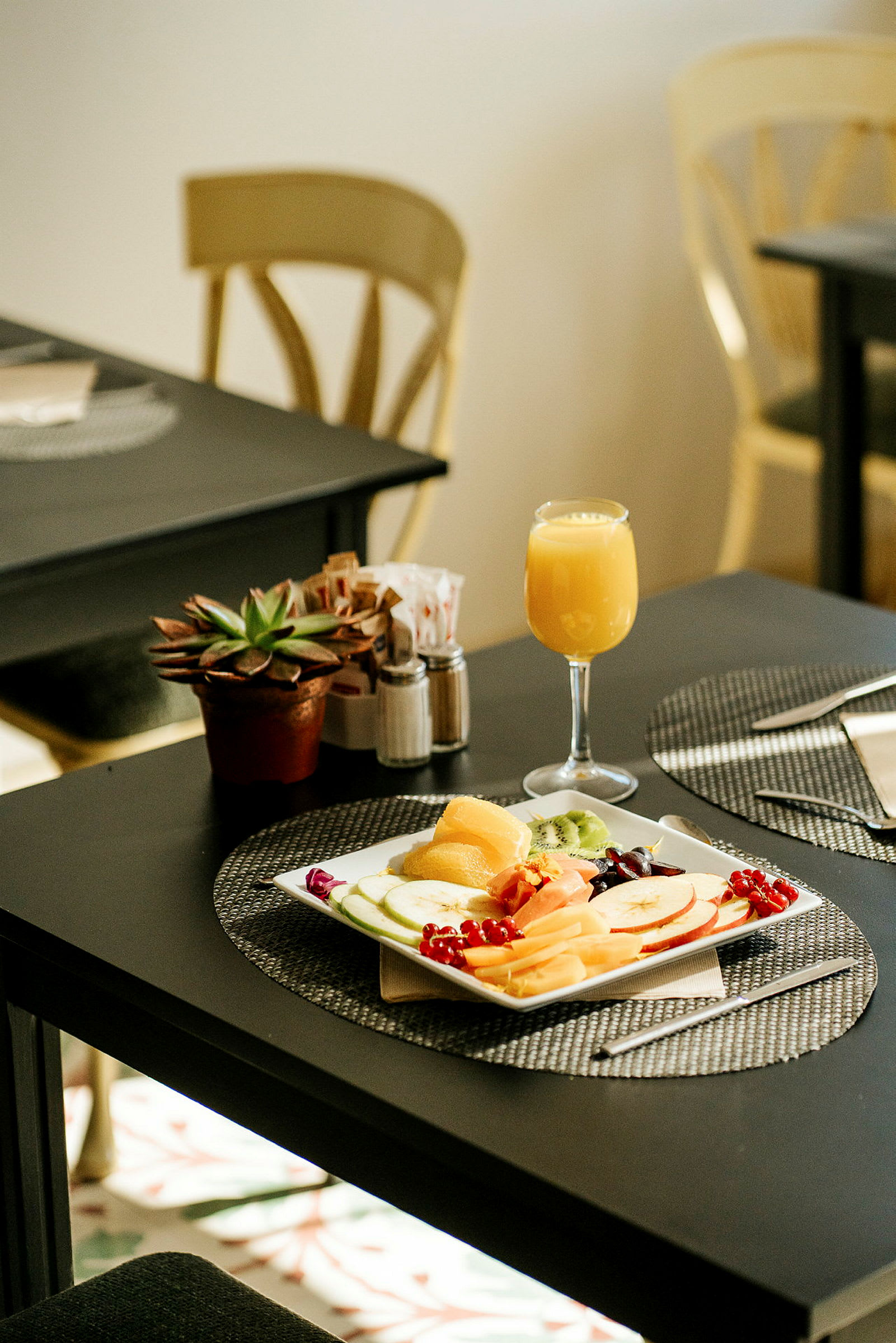 Ett bord med en tallrik fylld med frukt, ett glas apelsinjuice och en krukväxt. (AI-genererad text)