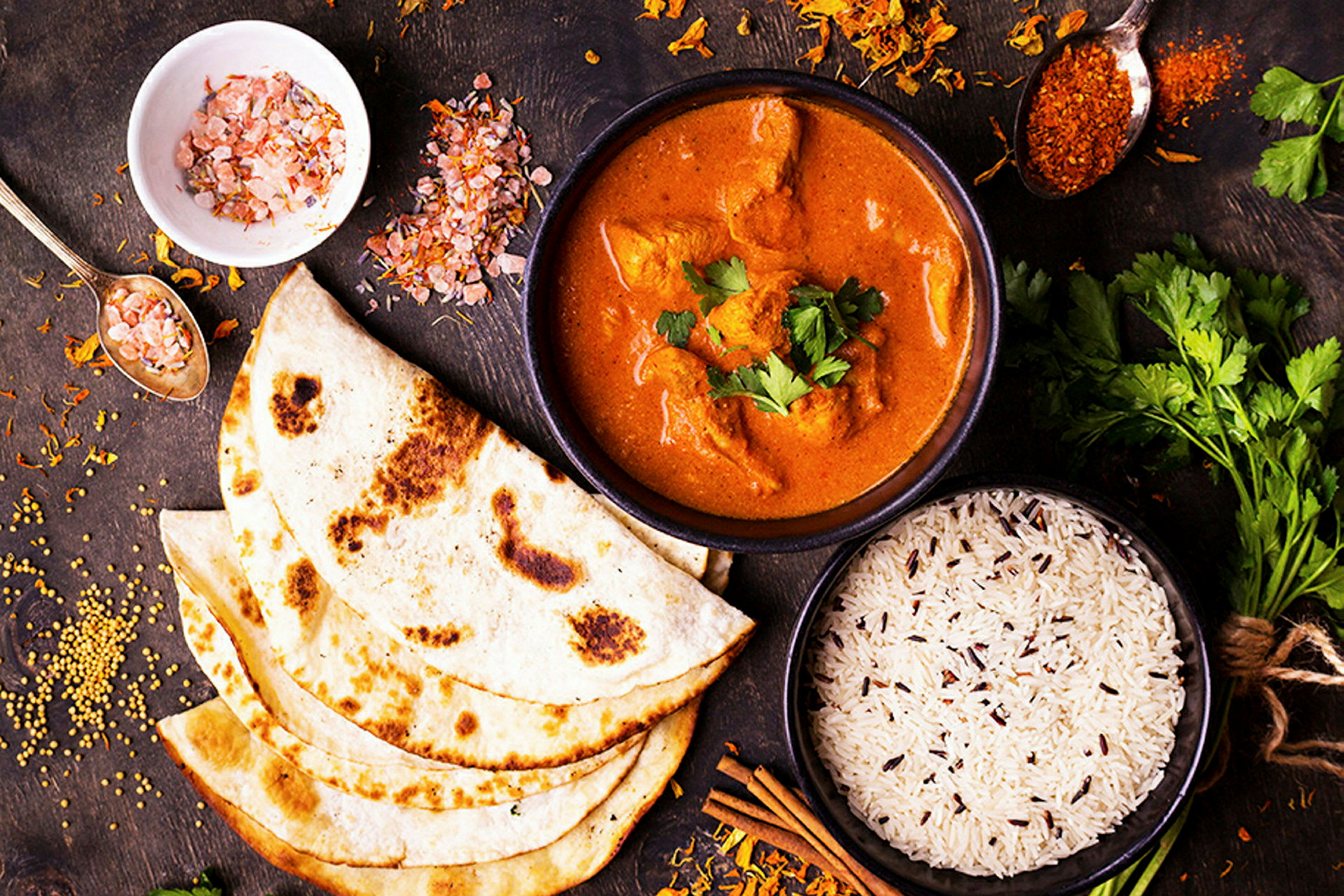 Ett upplägg med kryddad currygryta, ris och naanbröd, samt kryddor och örter på ett träbord. (AI-genererad text)