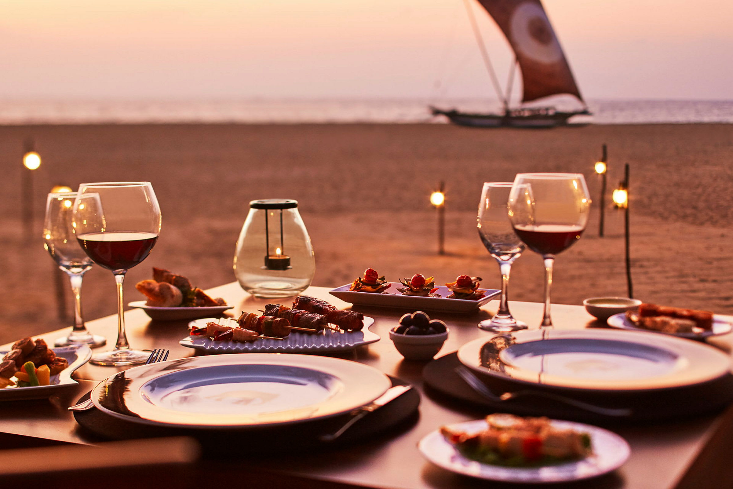 En dukad bord med mat och glas vin på stranden under solnedgången. (AI-genererad text)