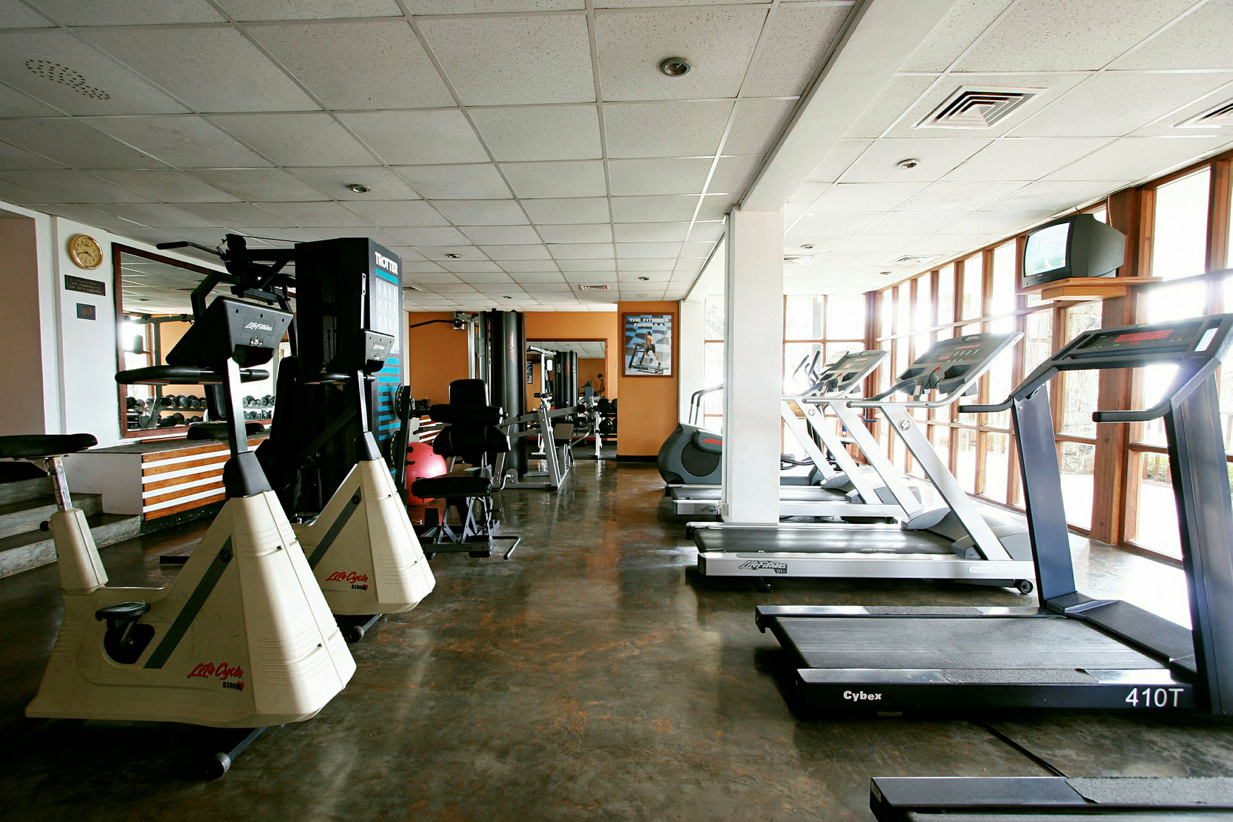 Ett gym med olika träningsmaskiner, inklusive löpband och motionscyklar, samt stora fönster och en spegelvägg. (AI-genererad text)