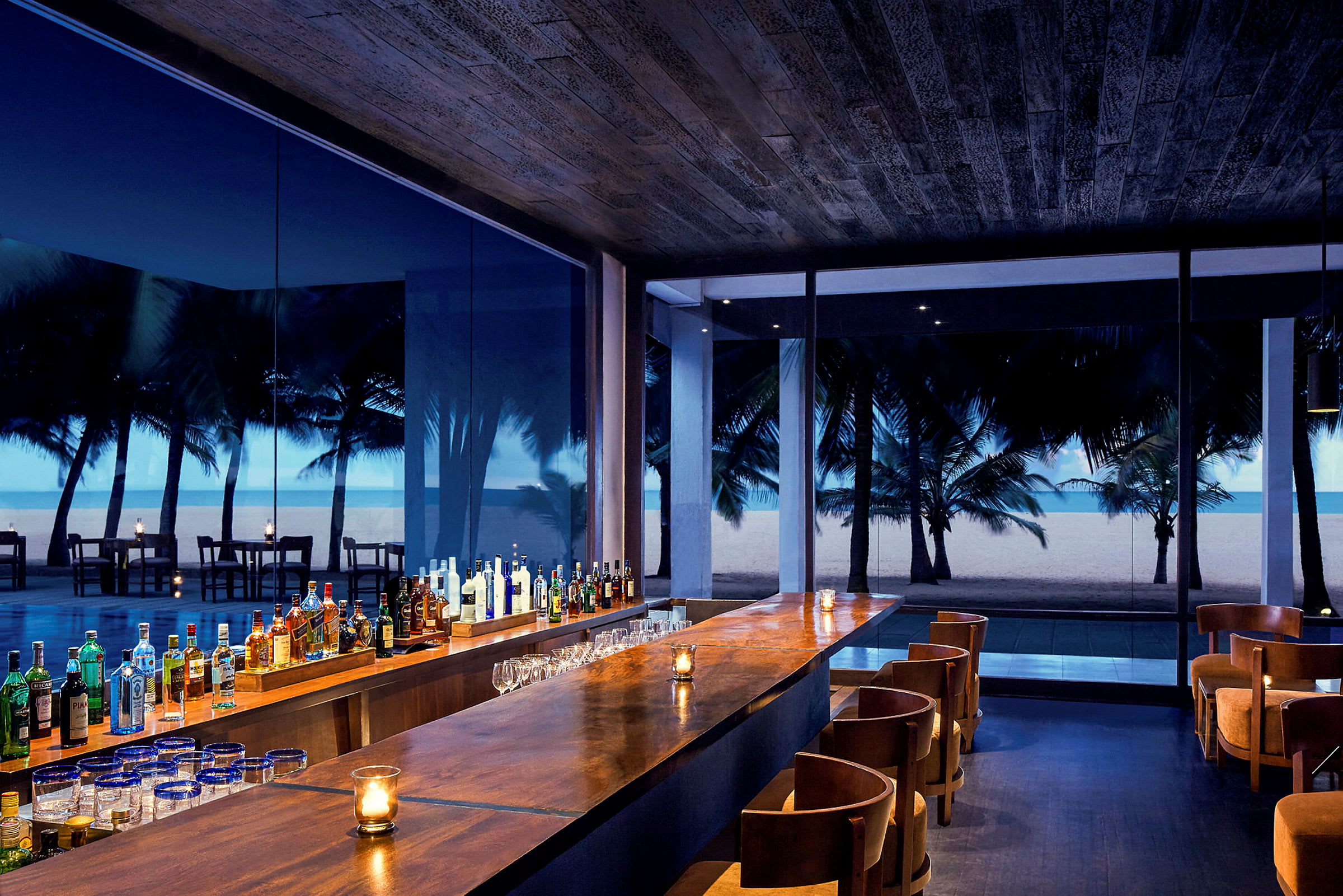 En bar med träinredning med utsikt över en sandstrand och palmer utanför. (AI-genererad text)