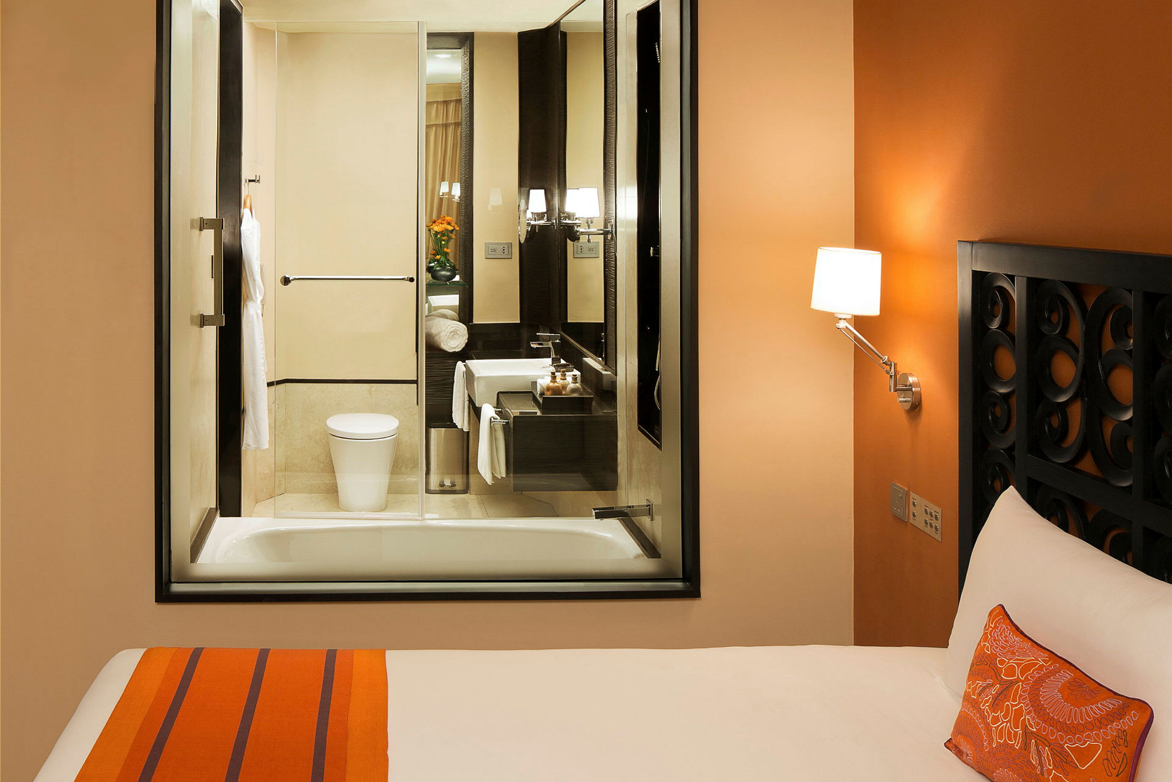 Hotellrum med orange inredning, säng och ett angränsande badrum med glasfönster. (AI-genererad text)