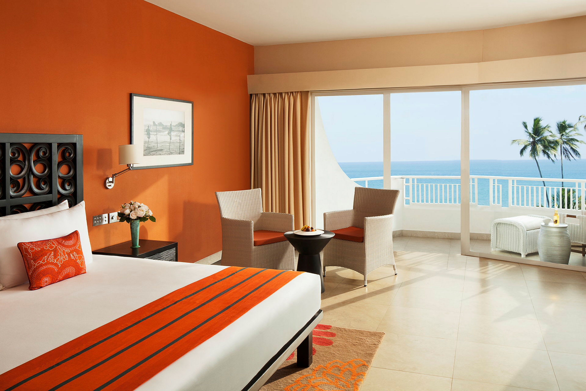 Ett hotellrum med orange väggar, en stor säng, två stolar vid ett bord och en balkong med utsikt över havet. (AI-genererad text)