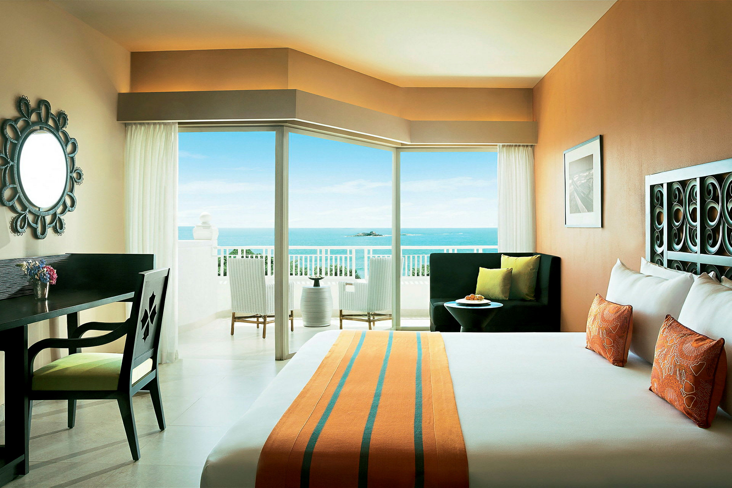 Ett hotellrum med en dubbelsäng, skrivbord, och en utsikt mot havet genom stora fönster och en balkong. (AI-genererad text)