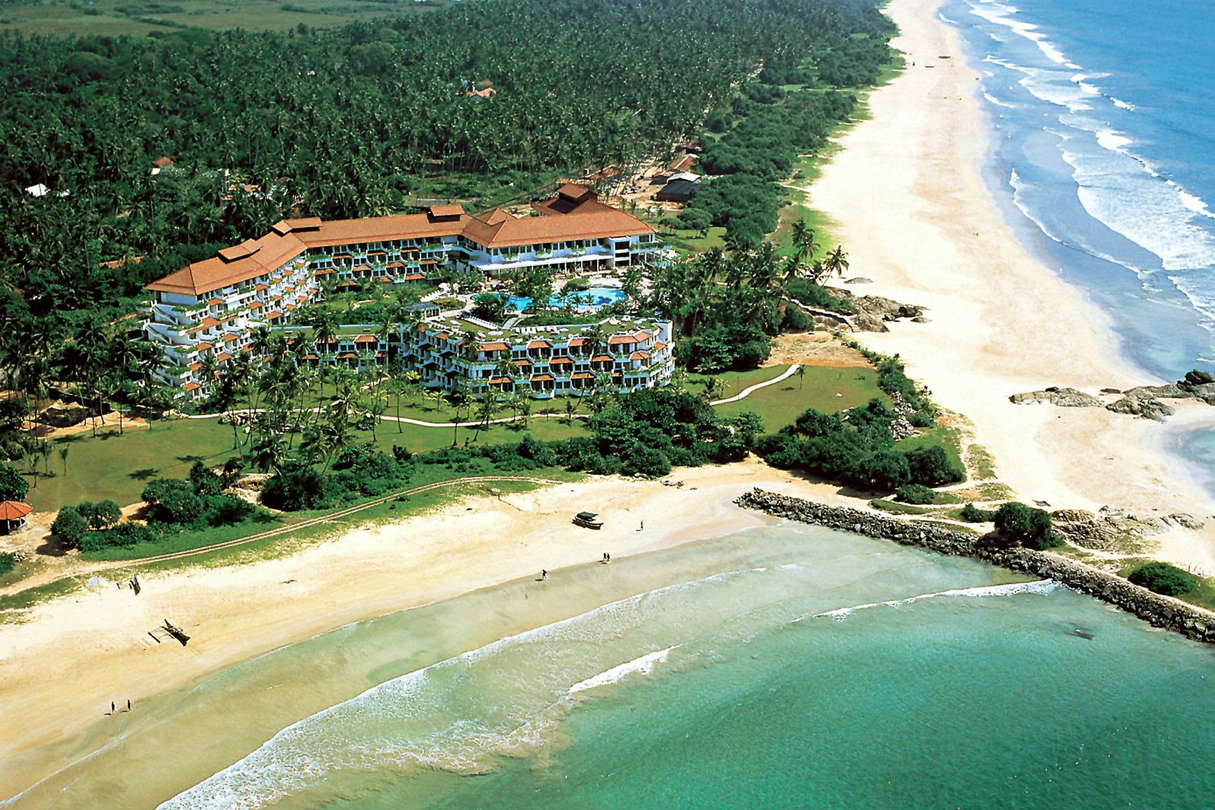 Flygbild av ett hotellkomplex omgivet av grönska vid kusten med en lång sandstrand och hav. (AI-genererad text)