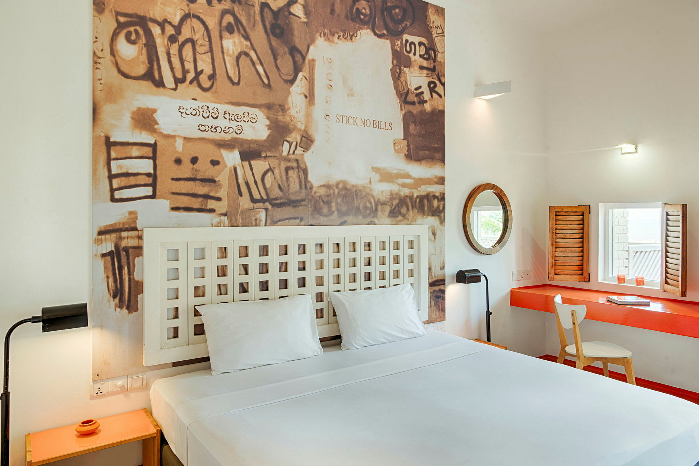 Ett hotellrum med en dubbelsäng, en tavla med graffiti-liknande konst på väggen bakom sängen och ett skrivbord med spegel och stol. (AI-genererad text)