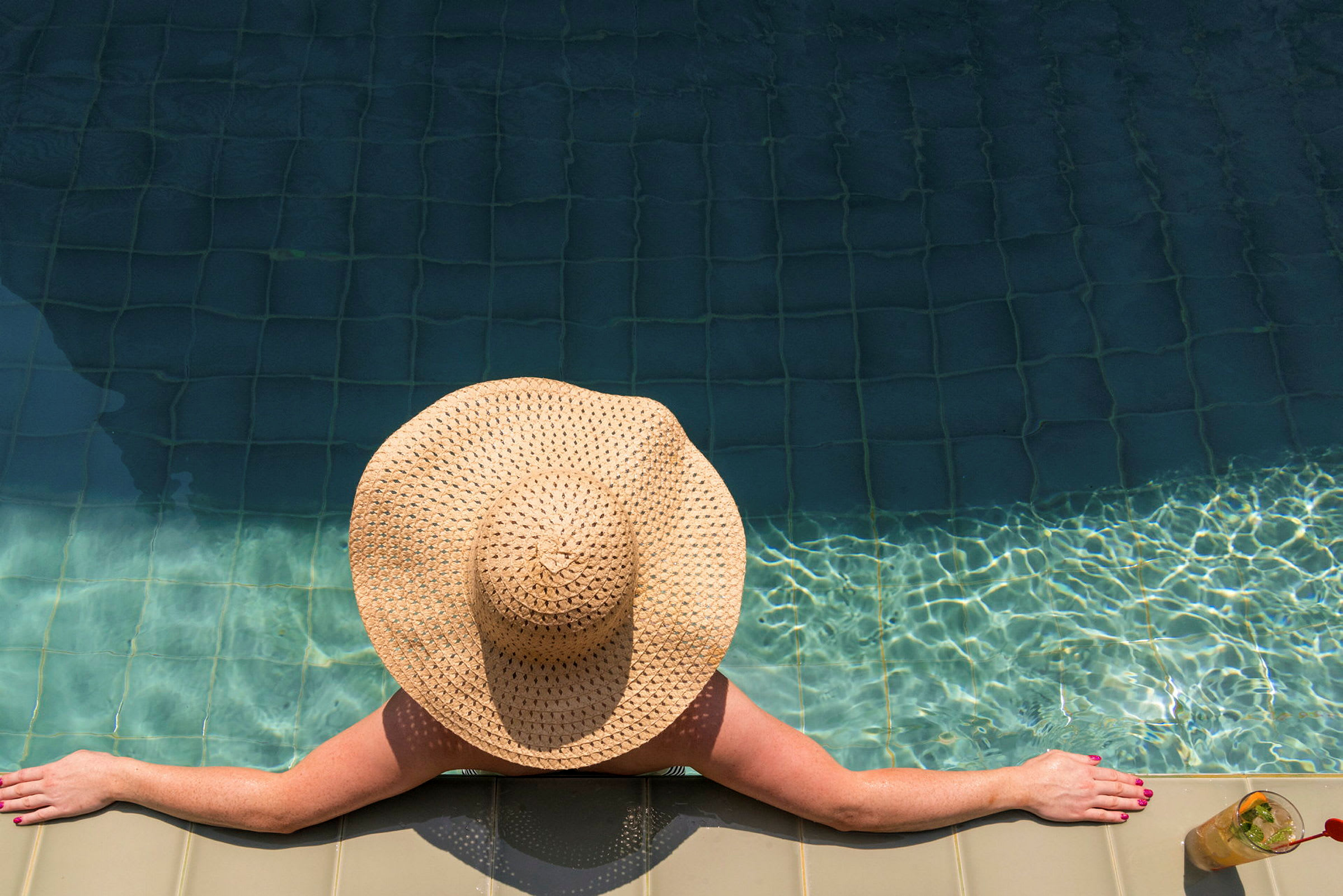 En person med en hattformad solhatt lutar sig mot kanten av en swimmingpool. (AI-genererad text)