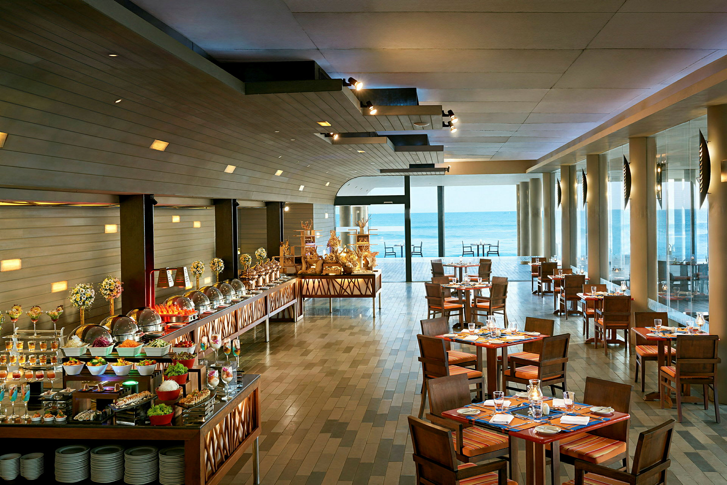 Bild av en restaurang med dukade bord och en buffé fylld med olika maträtter, samt stora fönster som vetter ut mot havet. (AI-genererad text)