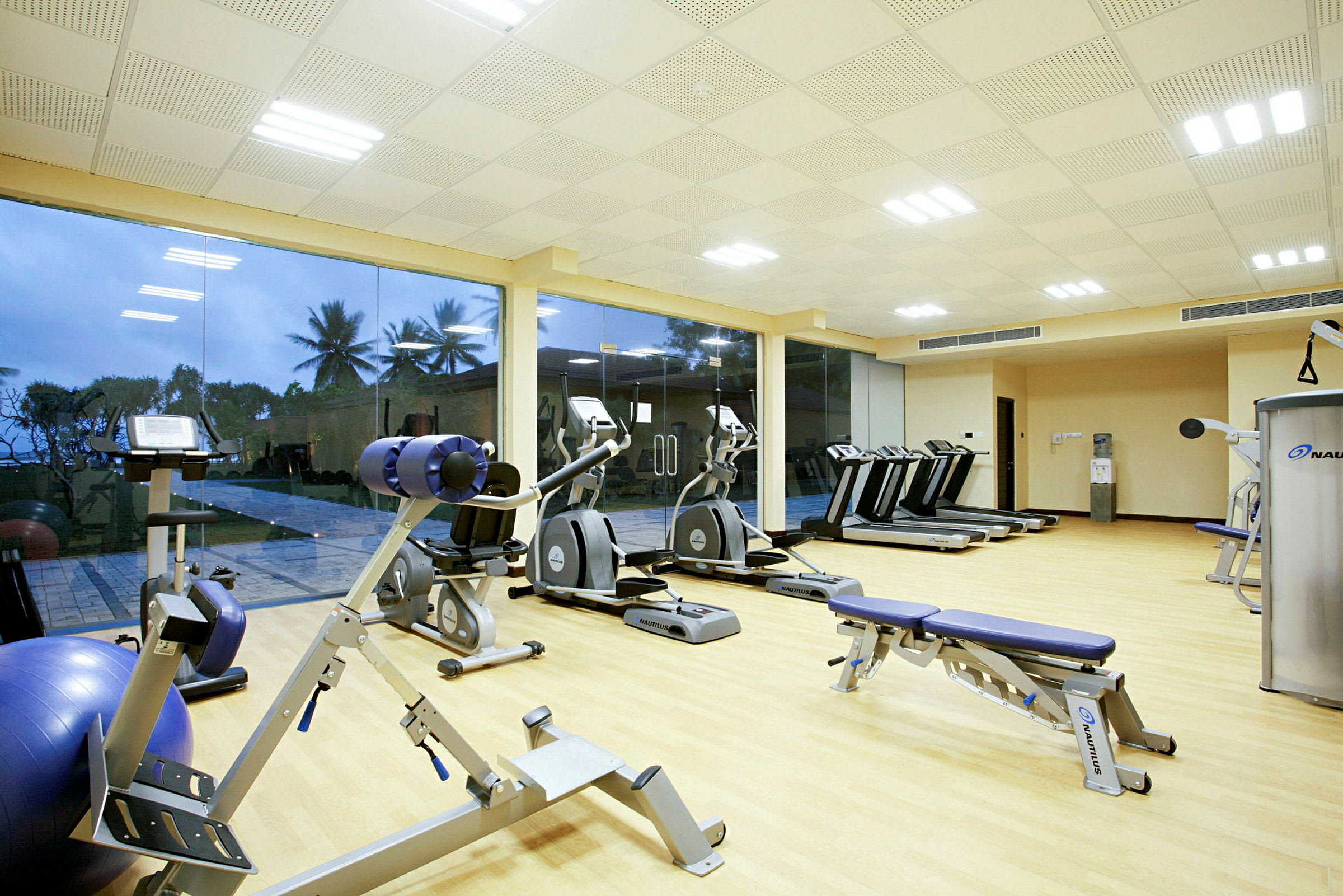 Ett gym med olika träningsmaskiner och fönster med utsikt mot palmer och utomhusmiljö. (AI-genererad text)
