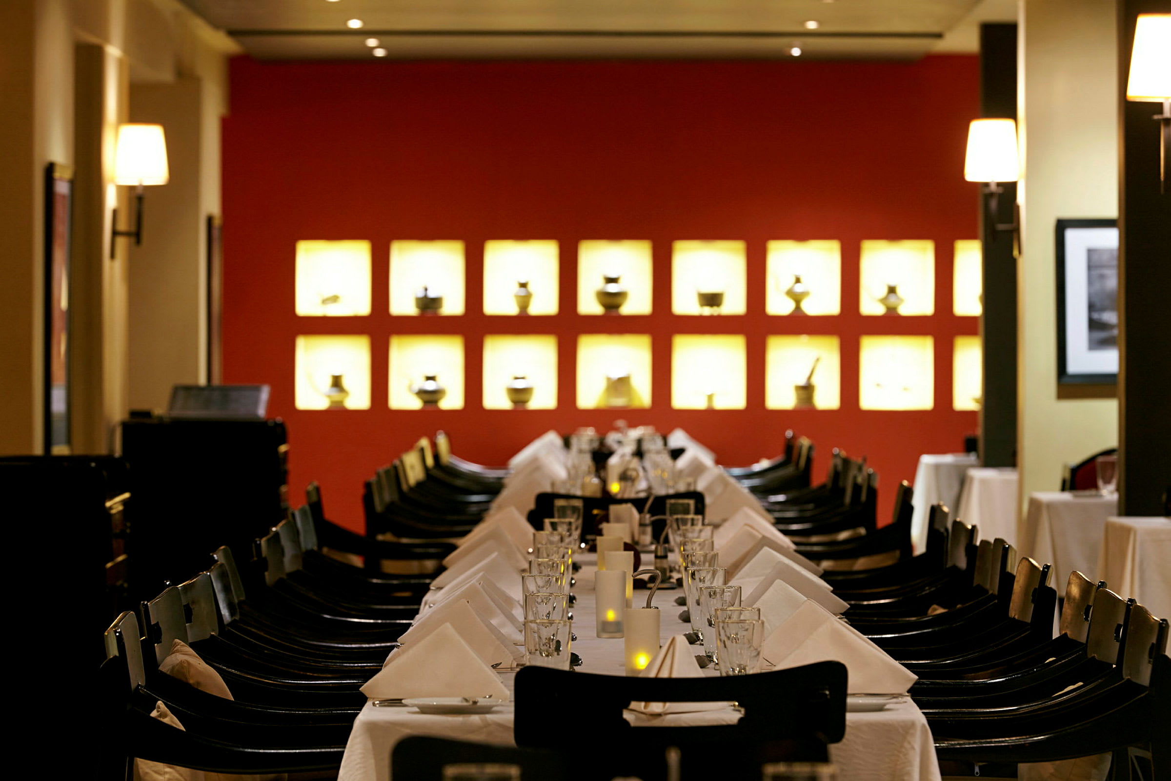 En restaurang med ett dukat bord täckt med vita dukar, glas och servetter. I bakgrunden finns en röd vägg med belysta små dekorativa föremål. (AI-genererad text)