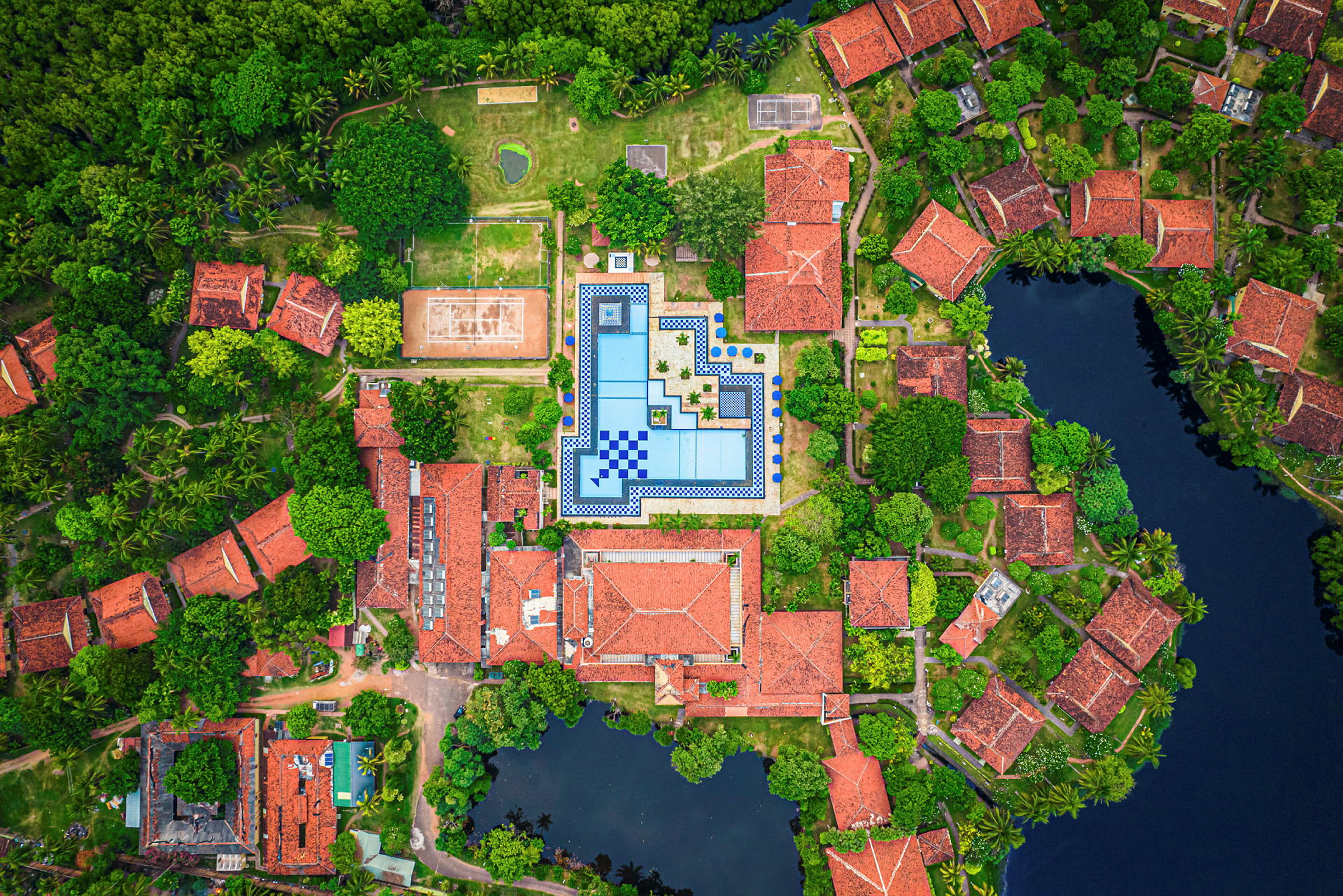 Färgstark överblick av hus med röda tak, en stor rektangulär pool och omgivande grönska. (AI-genererad text)