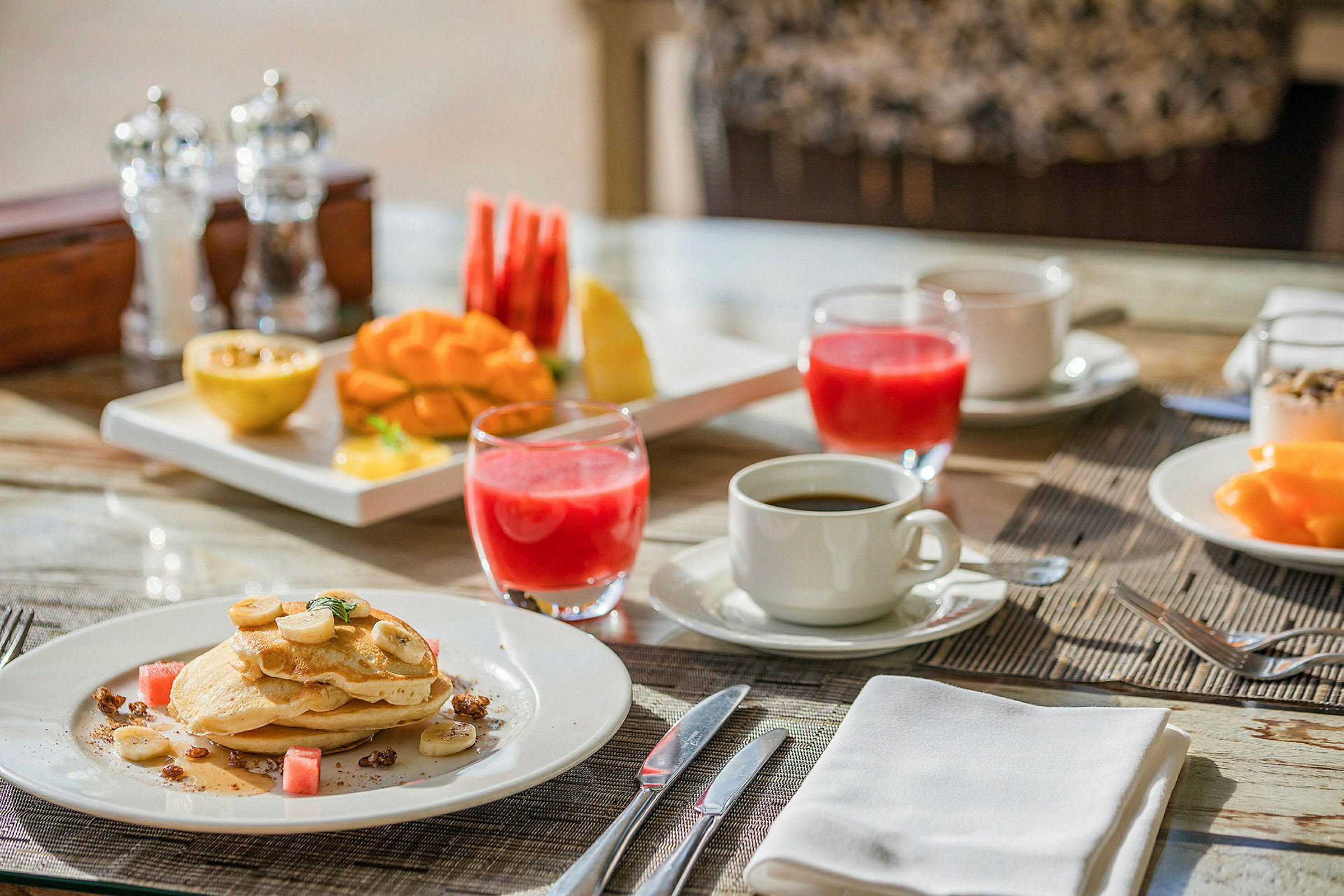 En frukostdukning med pannkakor, frukt, juice och kaffe på ett bord. (AI-genererad text)