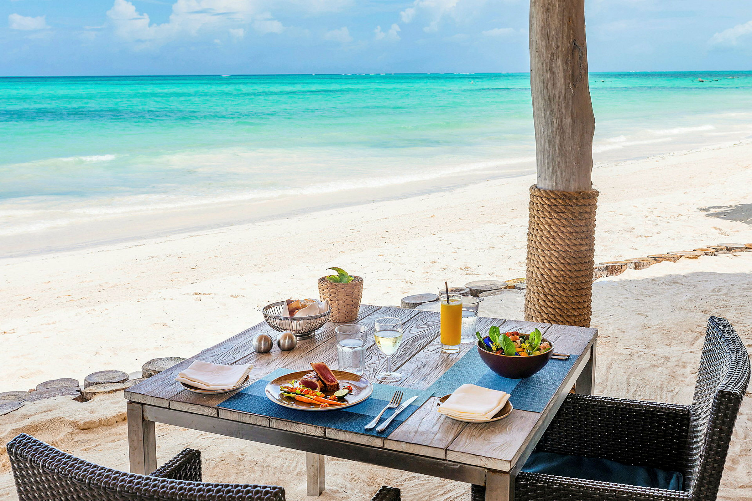 Ett dukat bord med mat och drycker på en sandig strand med havet i bakgrunden. (AI-genererad text)